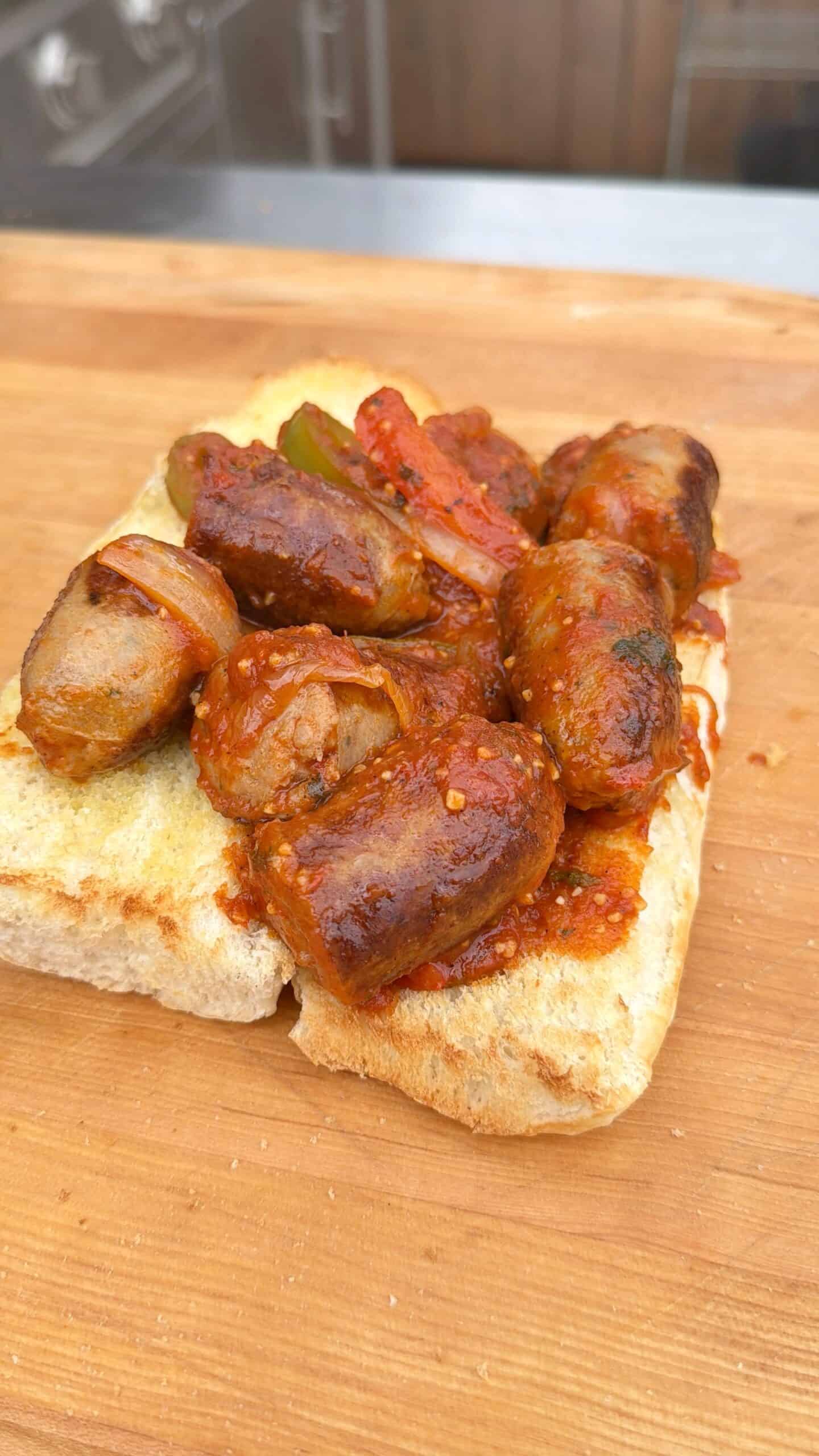 A piece of white bread topped with sausage and peppers pieces, bell peppers, and onions in a rich tomato sauce, served on a wooden surface.
