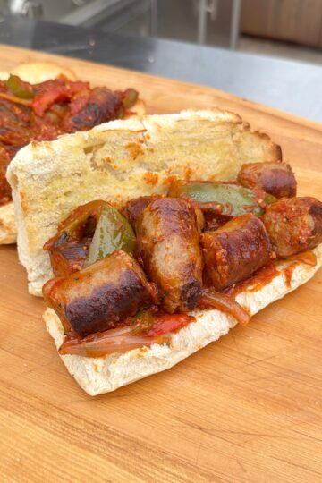 A toasted sandwich roll filled with grilled sausage slices, sautéed green peppers, and onions is placed on a wooden surface. Another similar sandwich is partially visible in the background.