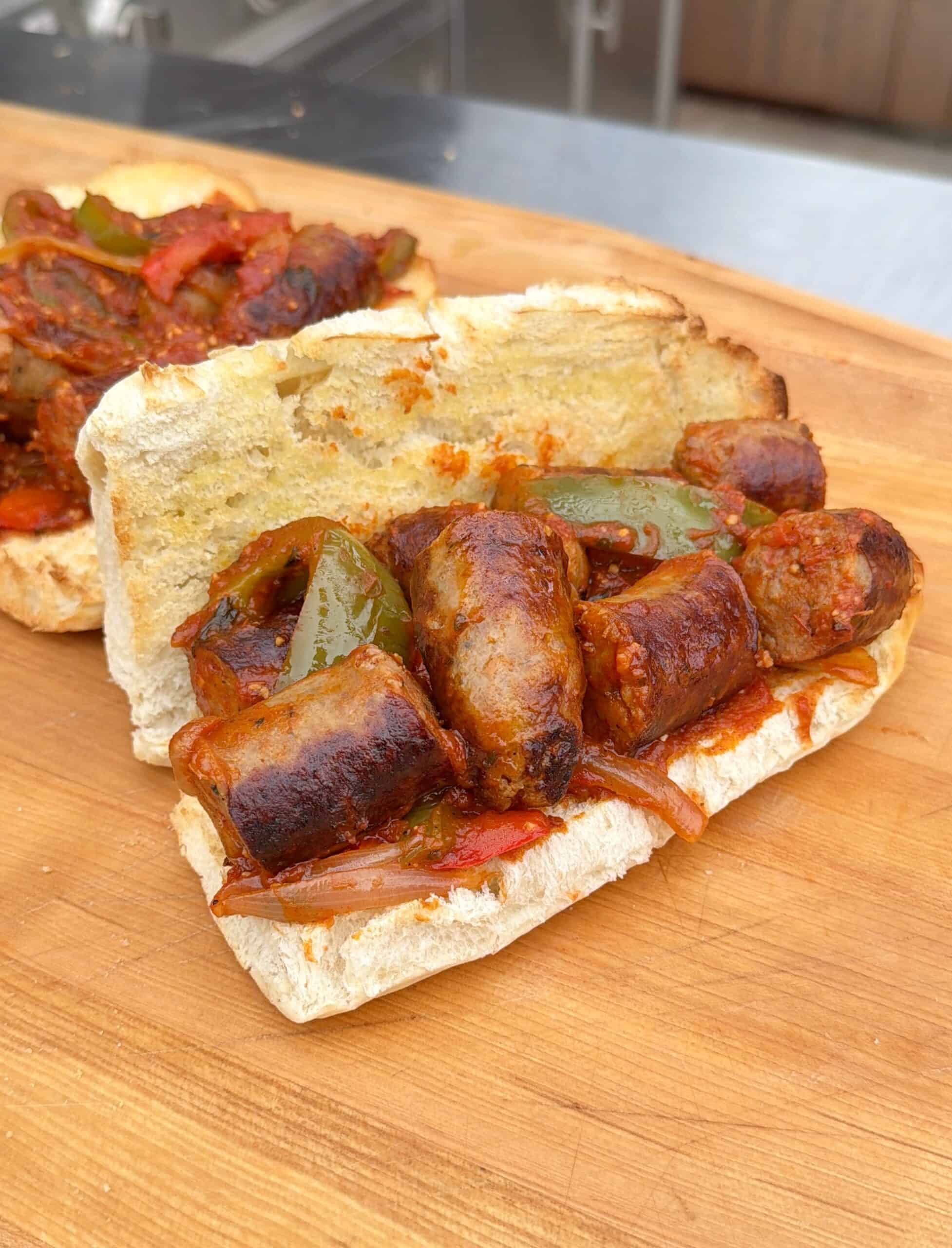 A toasted sandwich roll filled with grilled sausage slices, sautéed green peppers, and onions is placed on a wooden surface. Another similar sandwich is partially visible in the background.