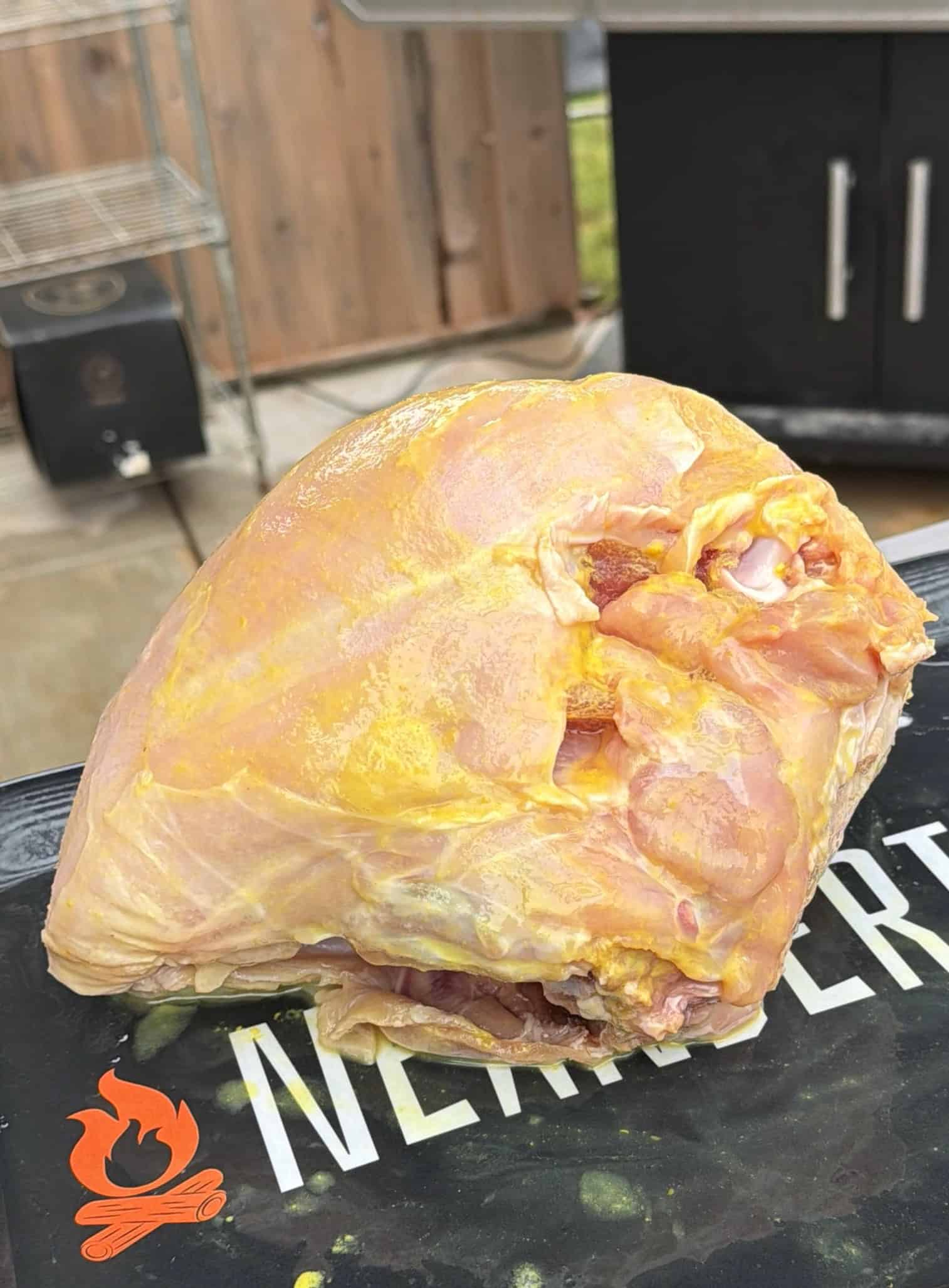 A large, raw turkey breast coated with Cajun seasoning sits on a tray outdoors, near a grill and smoker, ready to be cooked.