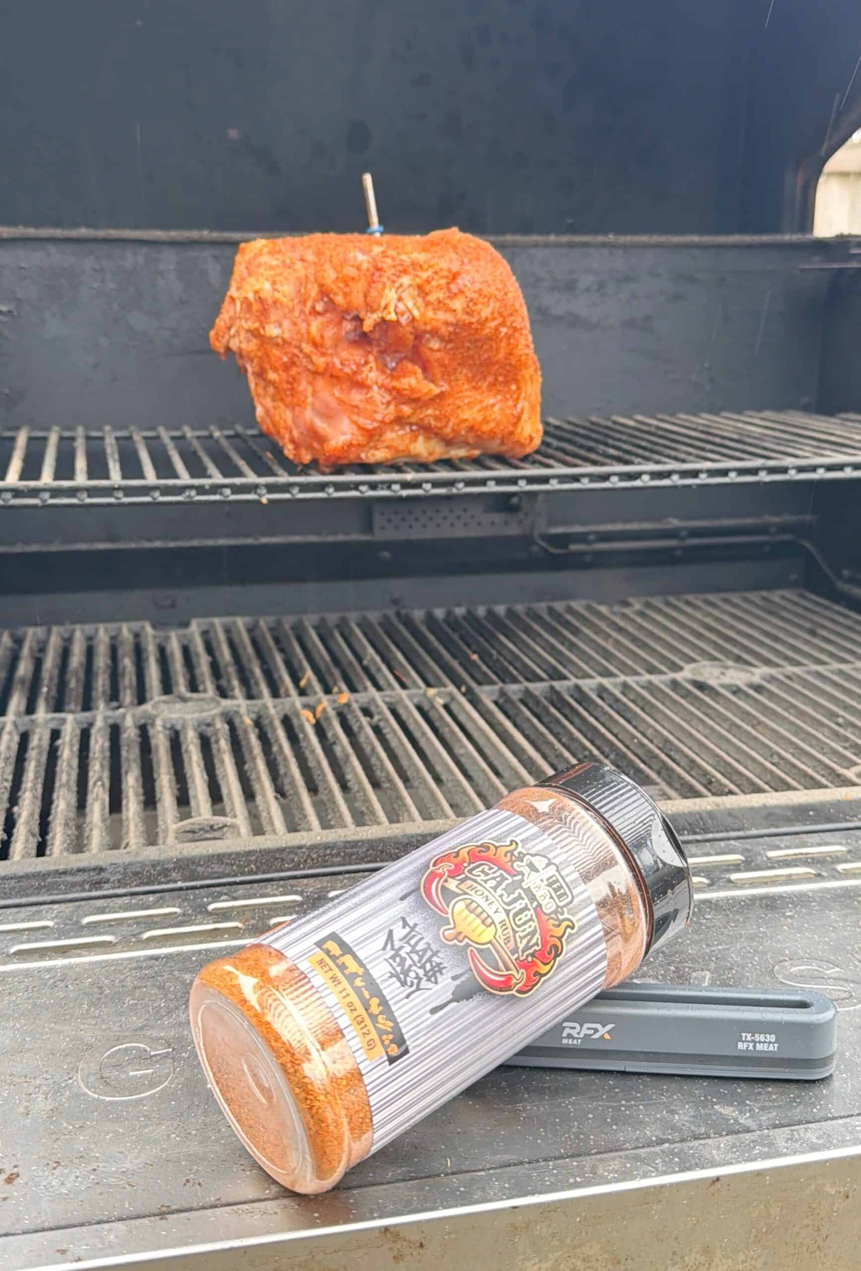 A seasoned pork roast sits on the upper rack of a barbecue grill. In the foreground, a container of Meat Church BBQ seasoning lies on its side near barbecue tools and a smoked Cajun turkey breast on the grill shelf.