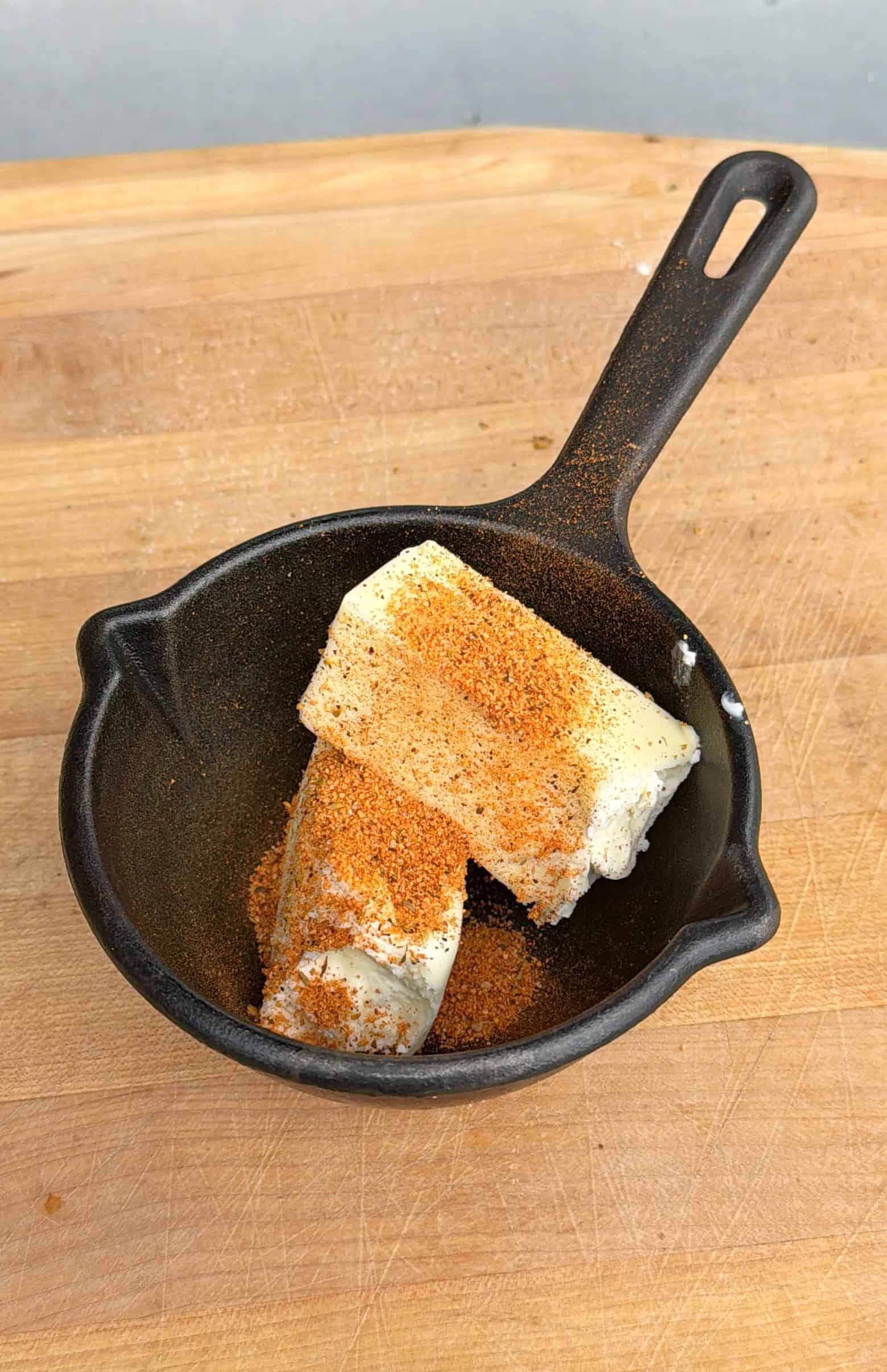 A small black cast iron saucepan on a wooden surface contains two blocks of cream cheese sprinkled with orange-brown Cajun turkey seasoning.