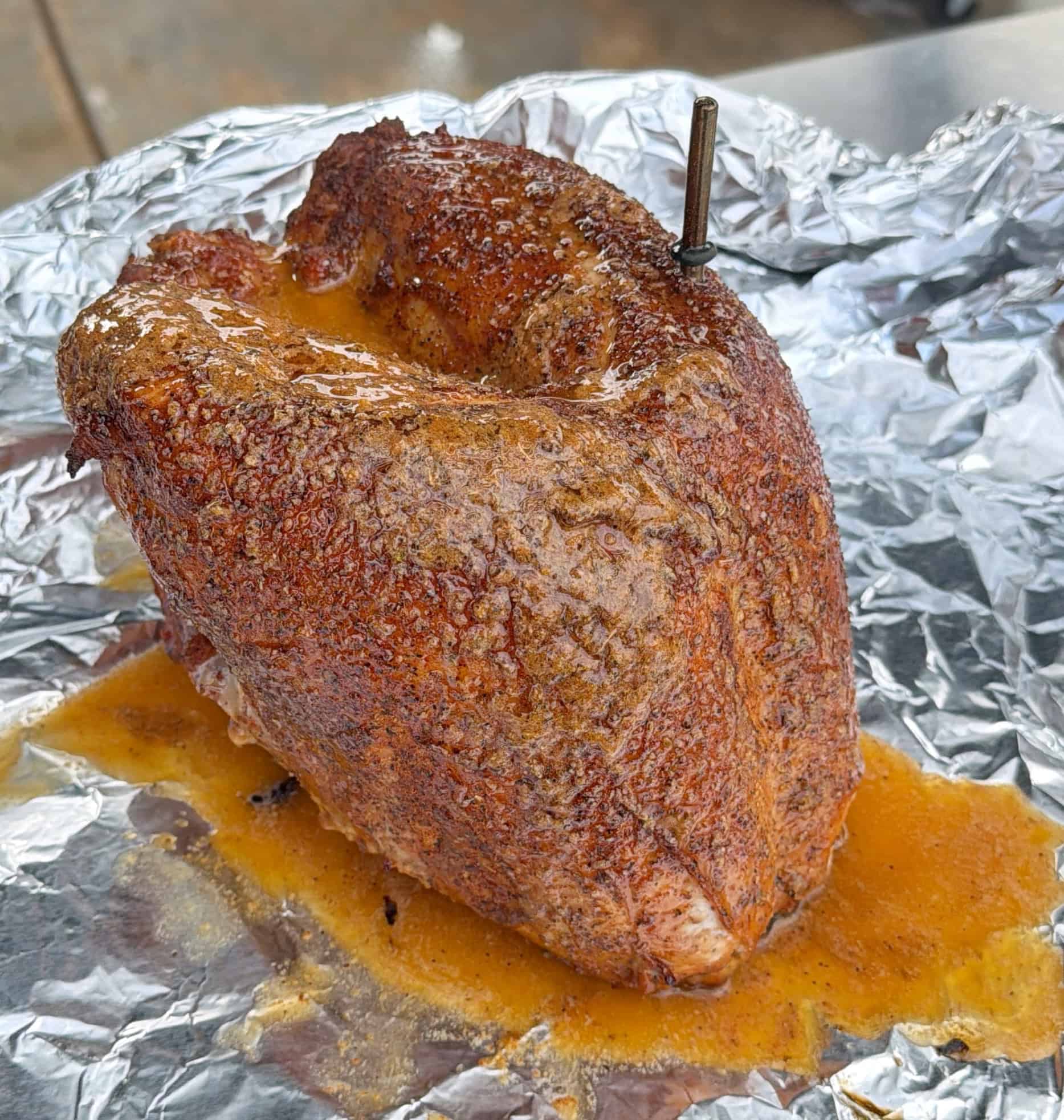A seasoned, juicy Honey Turkey Breast sits on crinkled aluminum foil, topped with a metal meat thermometer. Juices are pooling around the meat, indicating it has been recently cooked or smoked.