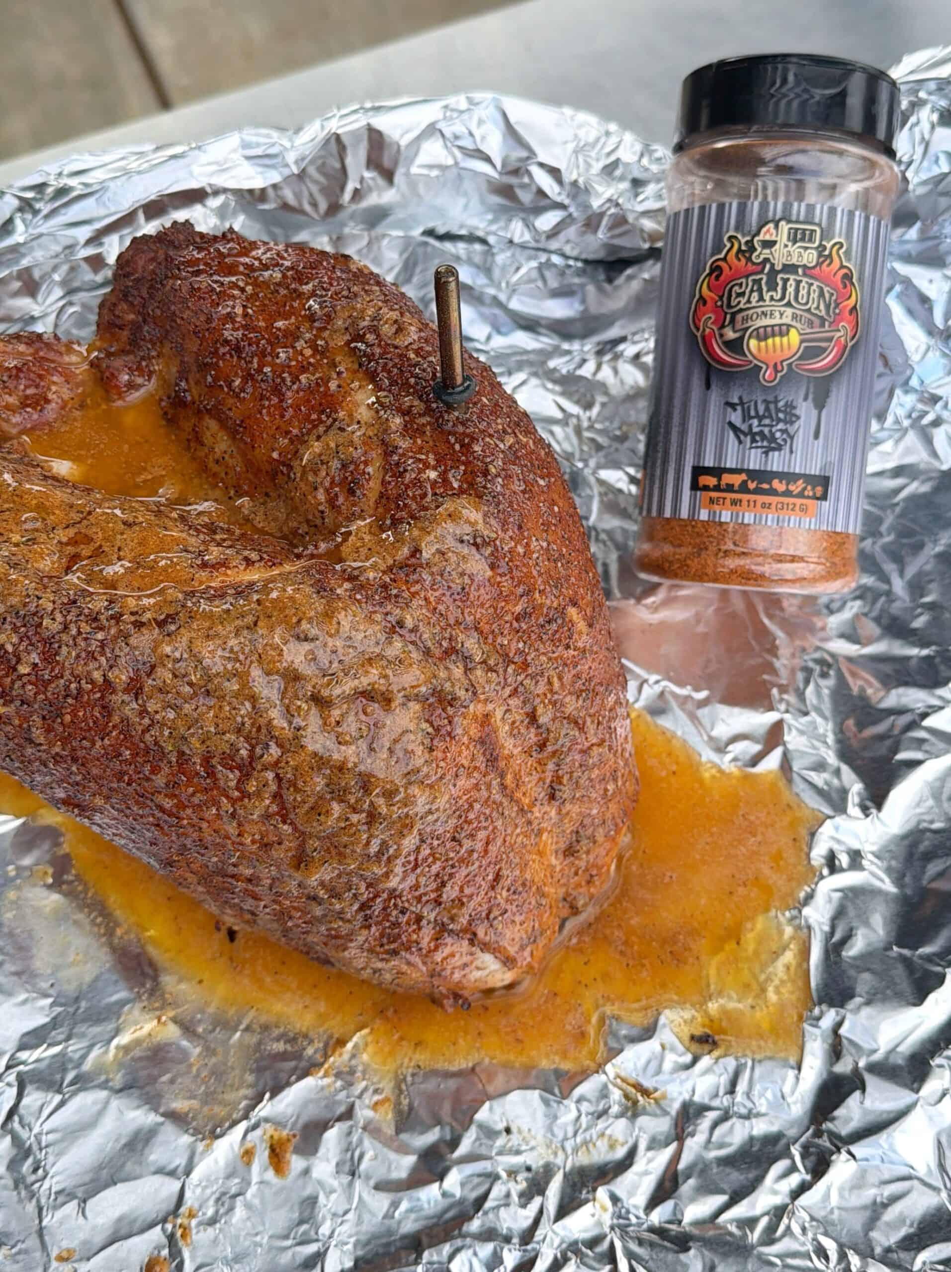 Seasoned, cooked Honey Turkey Breast resting on aluminum foil with a meat thermometer inserted, next to a bottle of Cajun seasoning blend. Juices are pooling around the turkey breast.