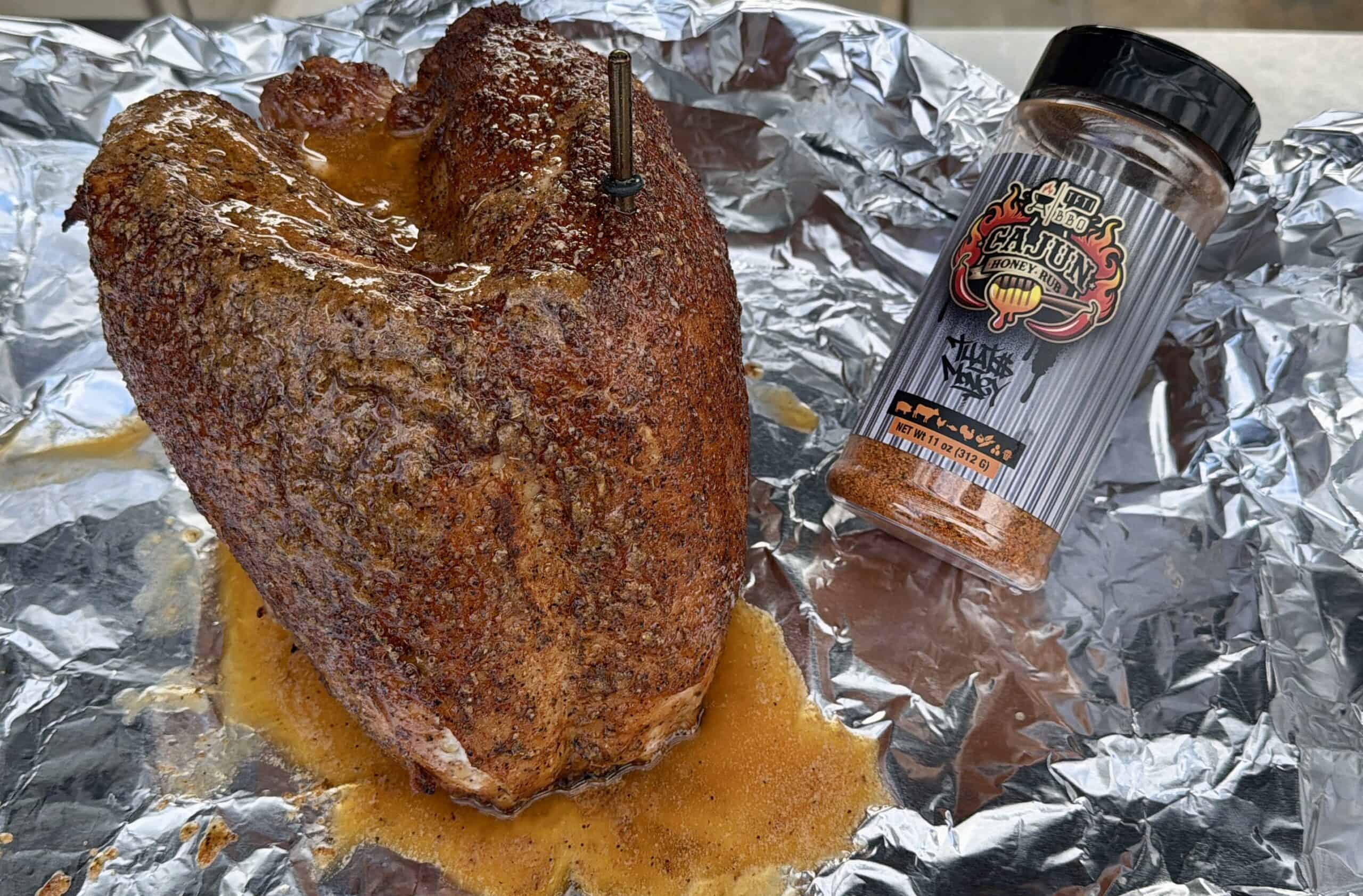 A seasoned, cooked Cajun honey Turkey Breast with a meat thermometer rests on foil, next to a spice bottle labeled Cajun Black Magic. Juices from the turkey breast pool on the foil.