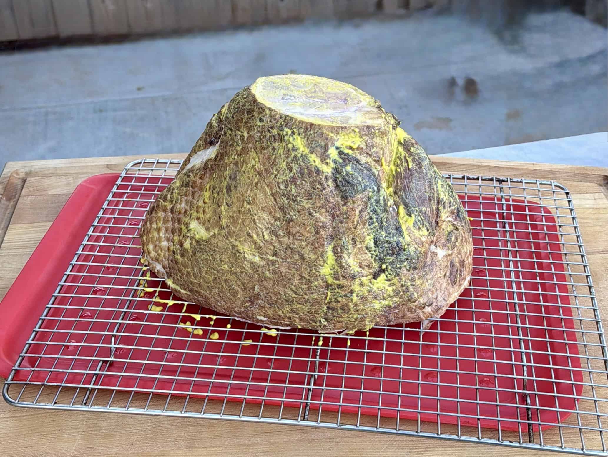 A large, uncooked ham coated with seasoning, resting on a metal cooling rack over a red tray on a wooden surface. The background is blurred, hinting at an outdoor setting.