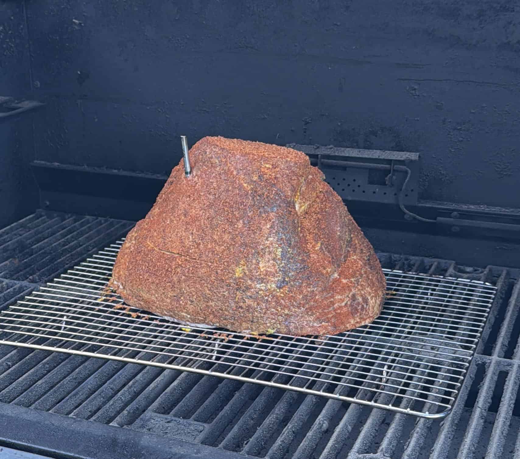 A large, seasoned brisket with a meat thermometer inserted is placed on a wire rack inside a smoker or grill, ready for slow cooking.