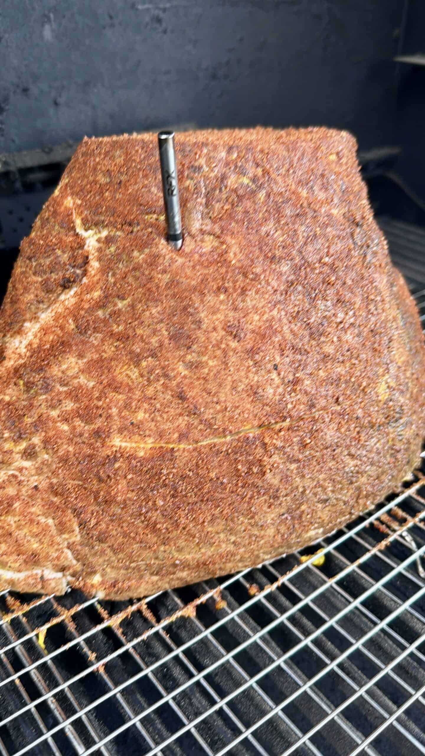 A large, seasoned cut of meat with a thermometer inserted sits on a metal rack inside a smoker, ready for cooking.