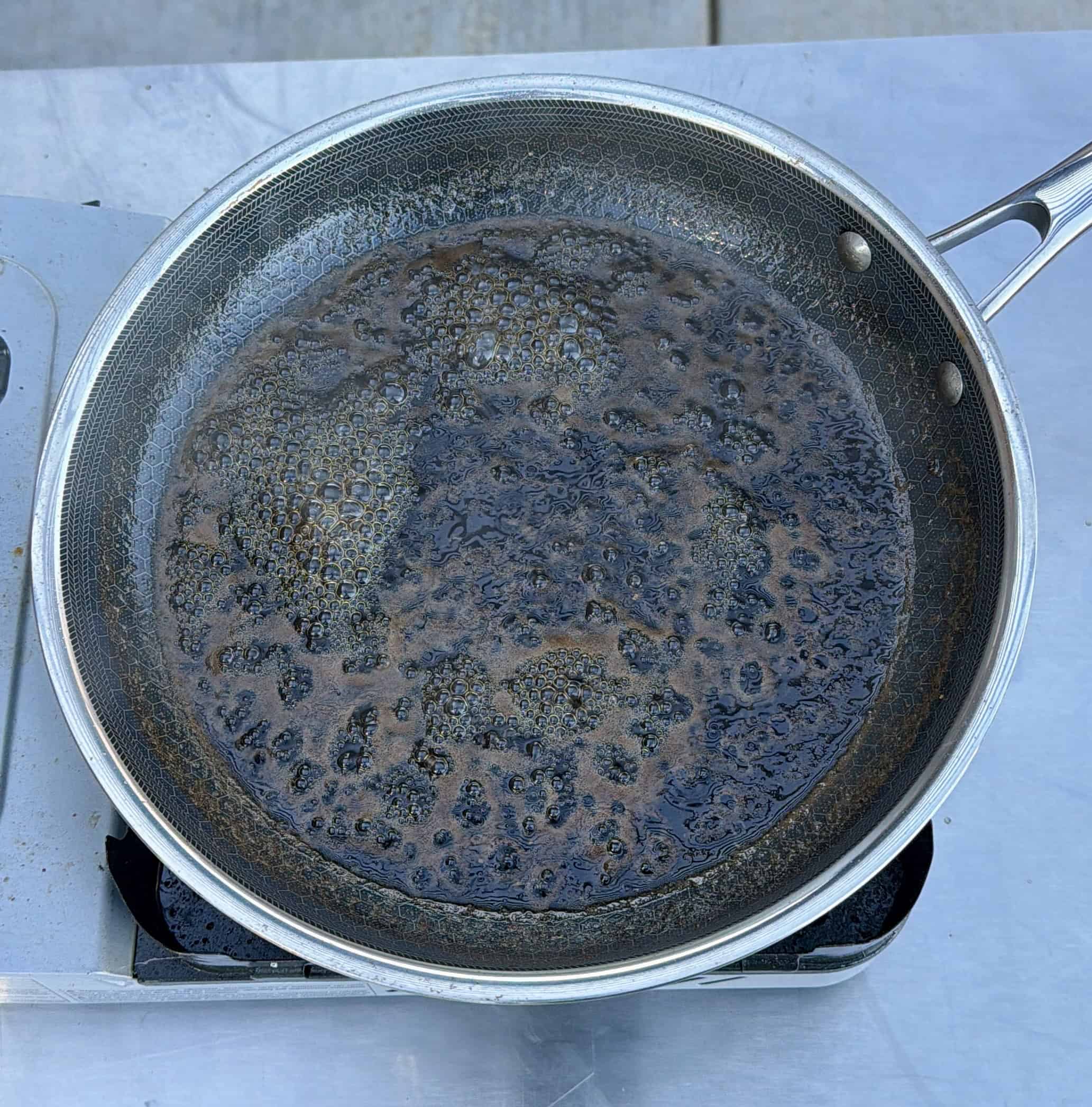 A stainless steel frying pan on a stove contains burnt, blackened residue and liquid, indicating overcooked or scorched food. The surface is uneven and charred.