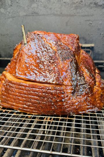 A glazed, spiral-cut ham with a meat thermometer inserted on top sits on a wire rack inside a smoker or grill. The ham has a shiny, caramelized coating and is being cooked slowly.