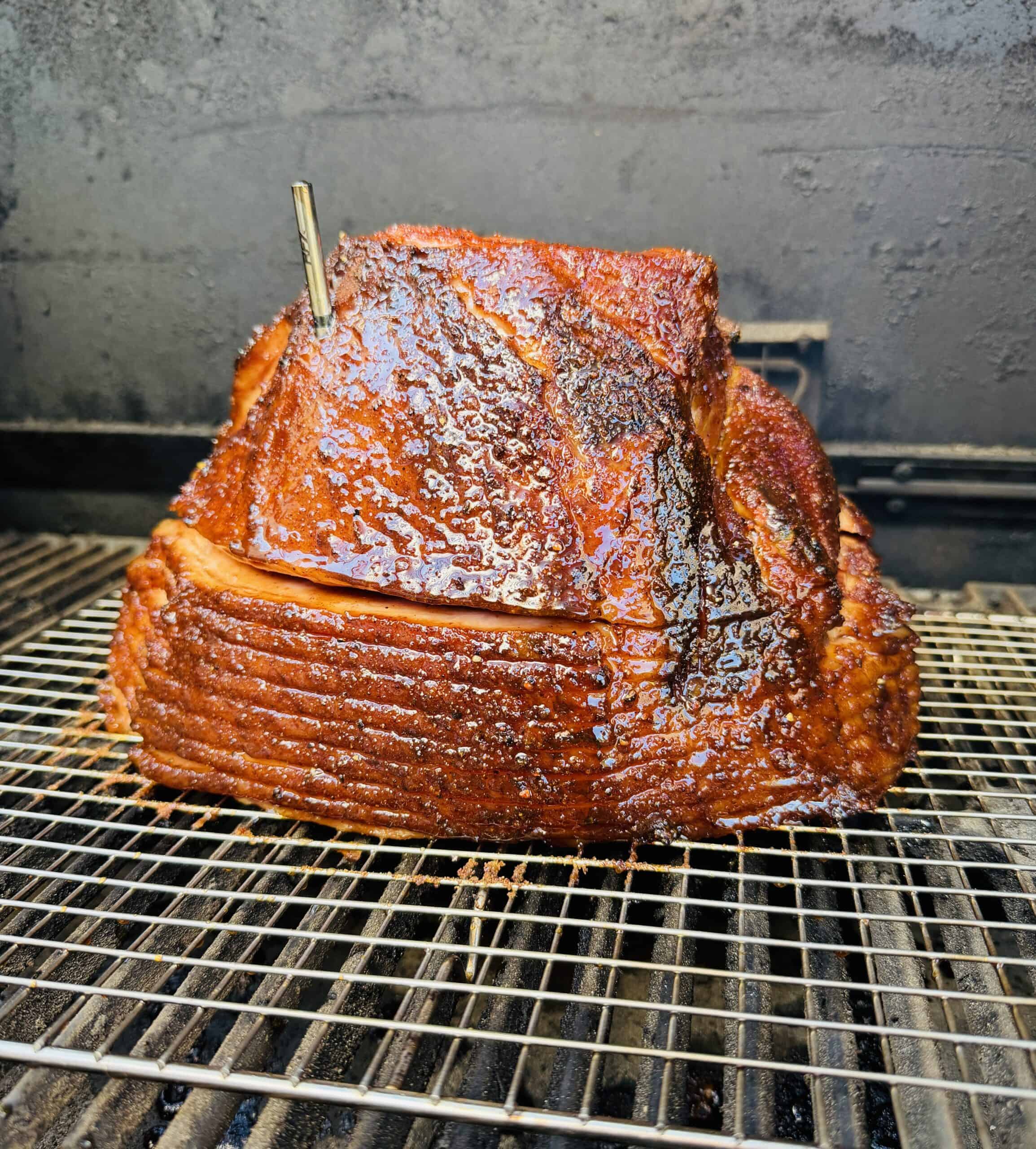 A dr pepper glazed ham, spiral-cut ham with a meat thermometer inserted on top sits on a wire rack inside a smoker or grill. The ham has a shiny, caramelized coating and is being cooked slowly.