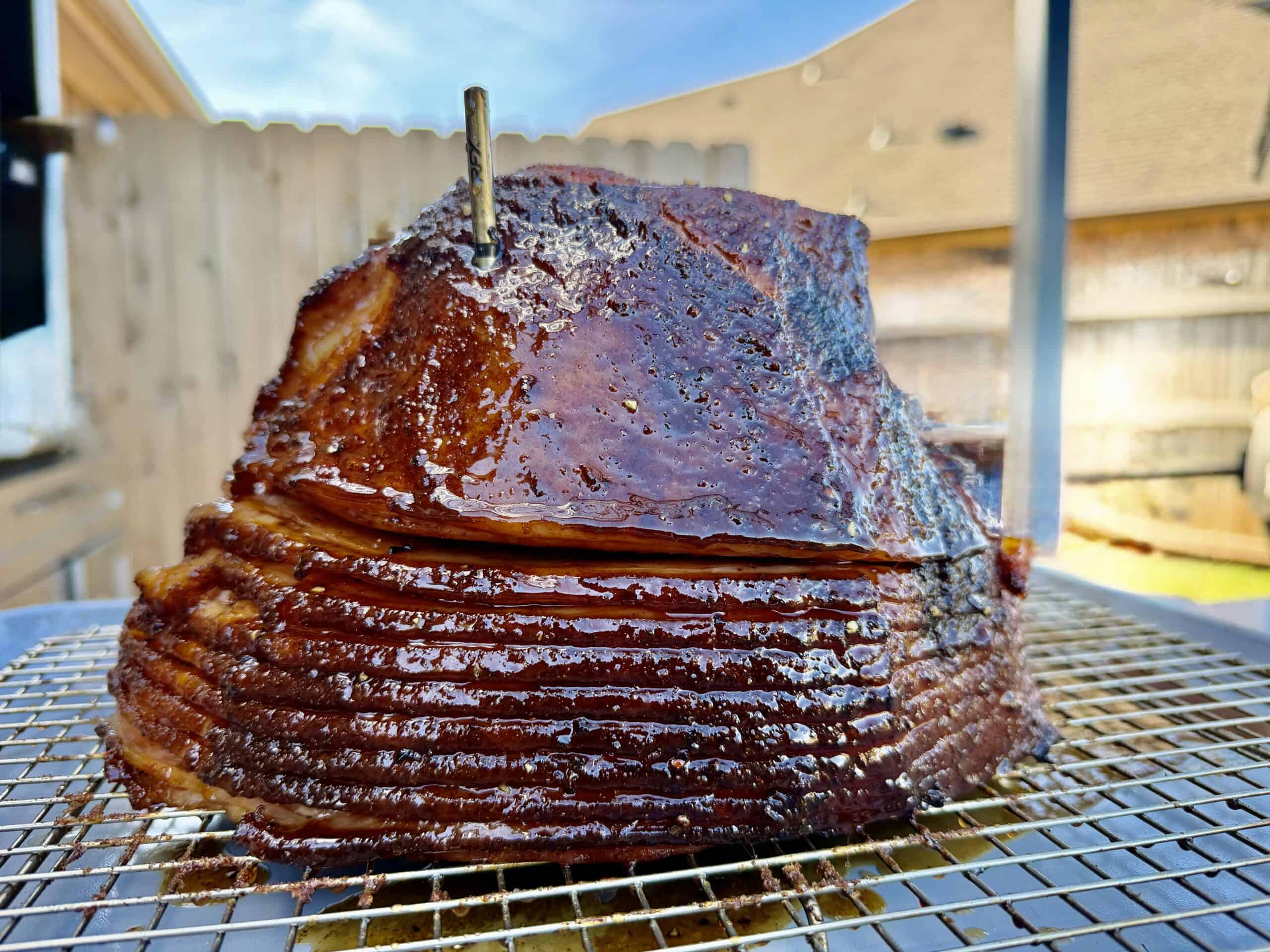 A close-up of a glazed, spiral-sliced ham resting on a wire rack outdoors, with a meat thermometer inserted on top. A wooden fence and neighboring houses are visible in the blurred background.