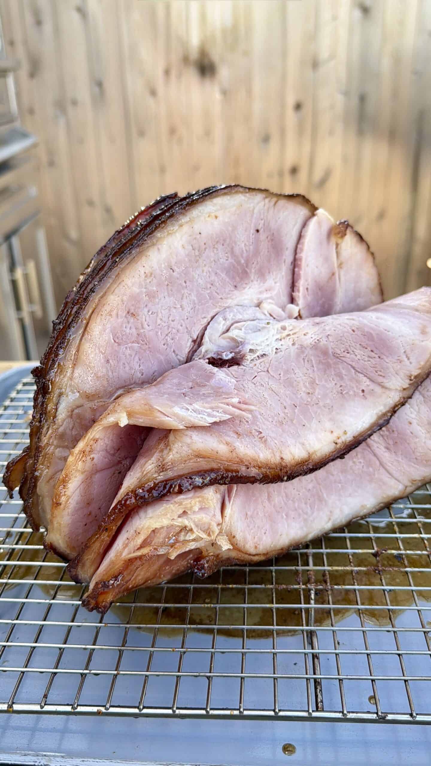 A large, spiral-cut ham rests on a wire rack, showcasing its pink, juicy interior and browned edges. The background appears to be an outdoor wooden fence.