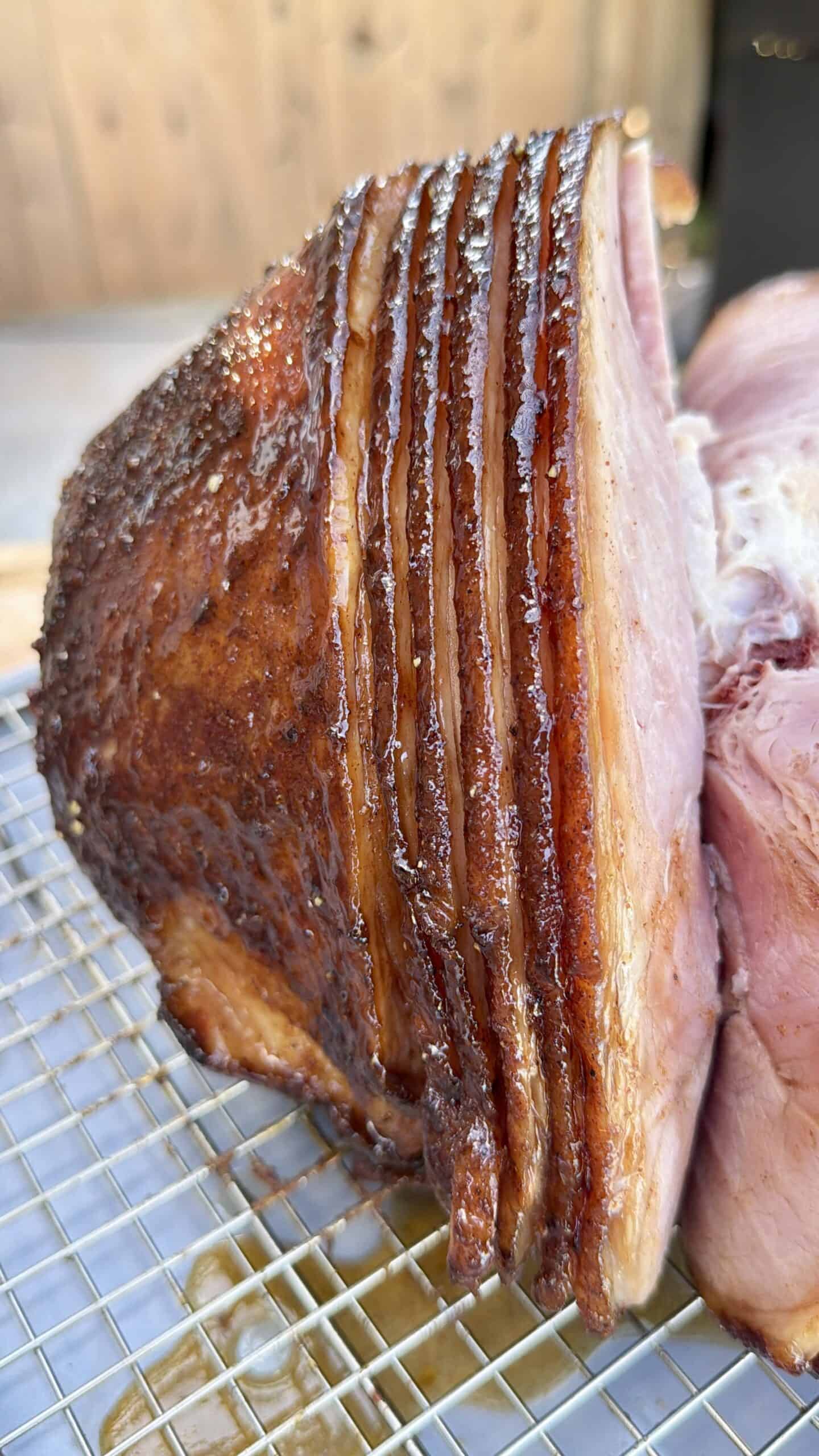 A close-up of a spiral-cut, glazed ham resting on a wire rack. The ham has a caramelized, golden-brown crust and juicy, pink interior with visible glaze and juices.