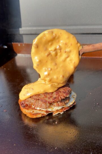 A thick, creamy cheese sauce with bits is being poured from a wooden spatula onto a grilled burger patty on a flat grill.