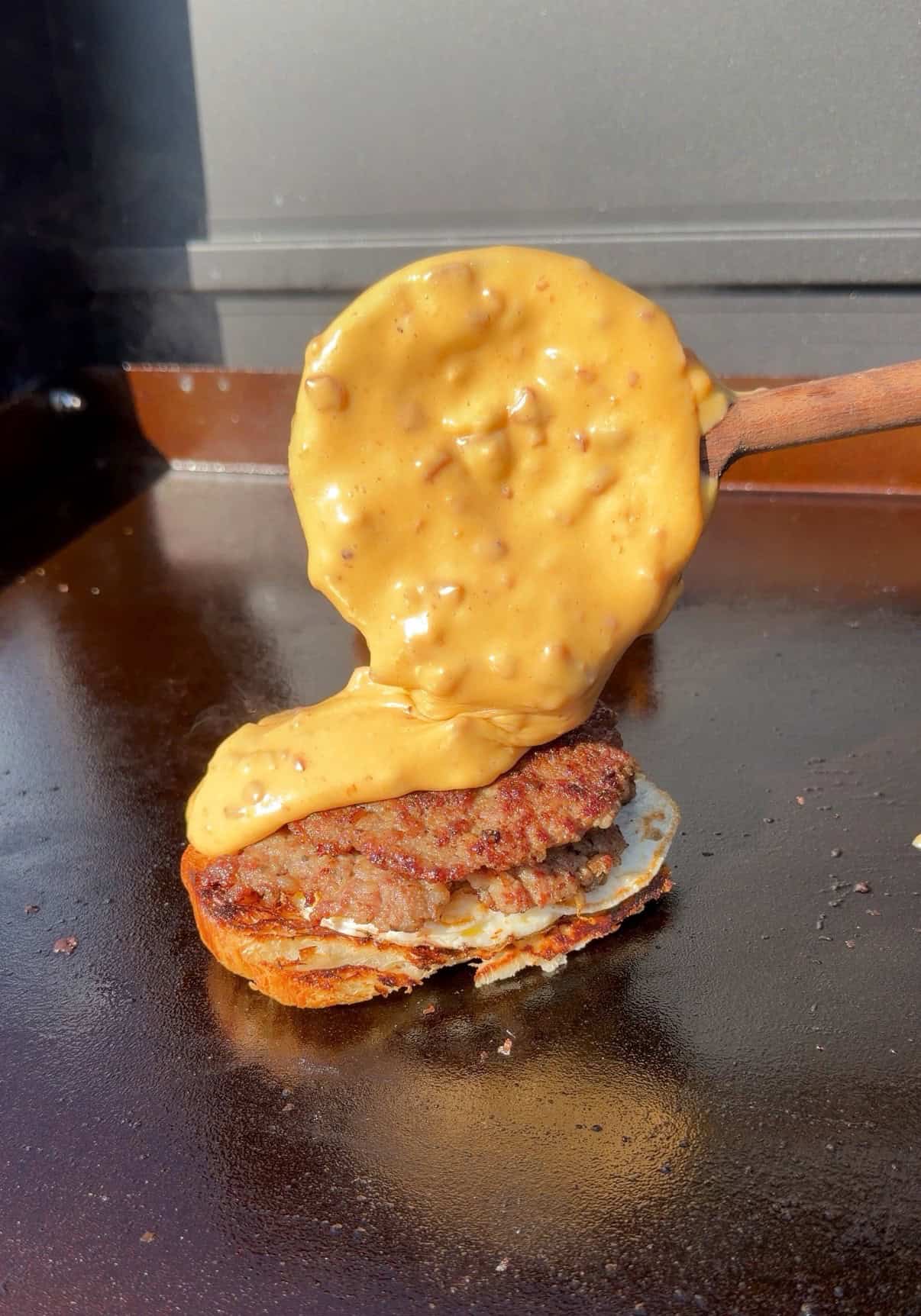 A thick, creamy cheese sauce with bits is being poured from a wooden spatula onto a grilled burger patty on a flat grill.