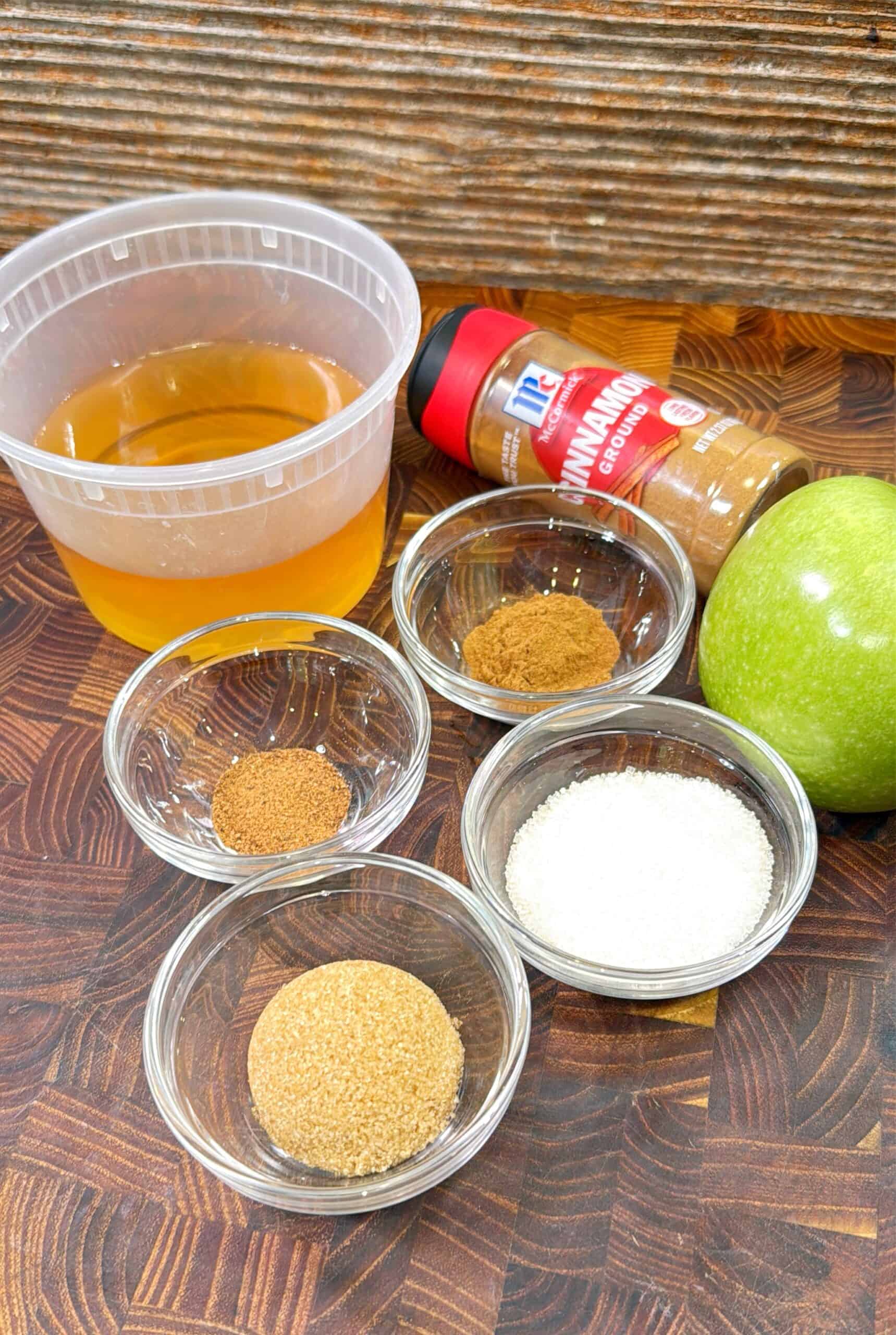 A plastic container of apple juice, a bottle of ground cinnamon, a green apple, and four small glass bowls containing brown sugar, white sugar, cinnamon, and nutmeg on a wooden surface.