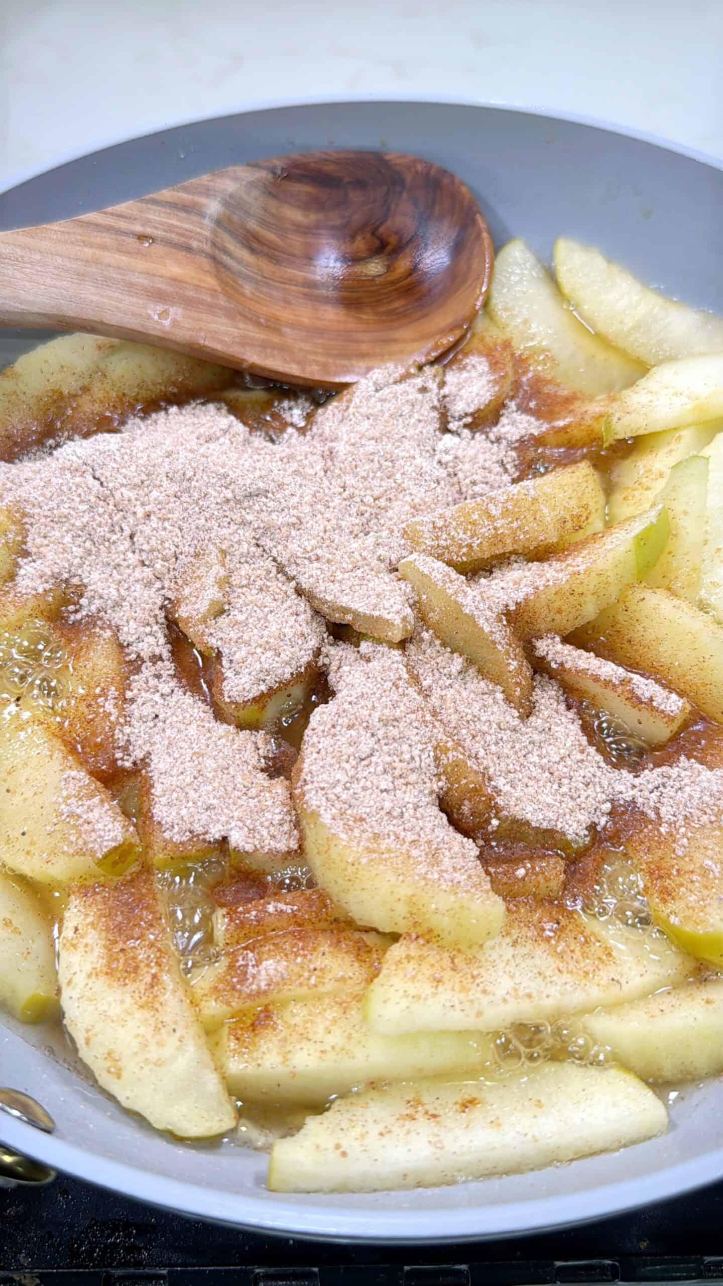 Apple slices are cooking in a pan with bubbling liquid, topped with a generous layer of cinnamon sugar. A wooden spoon rests on the side, ready to stir the mixture.