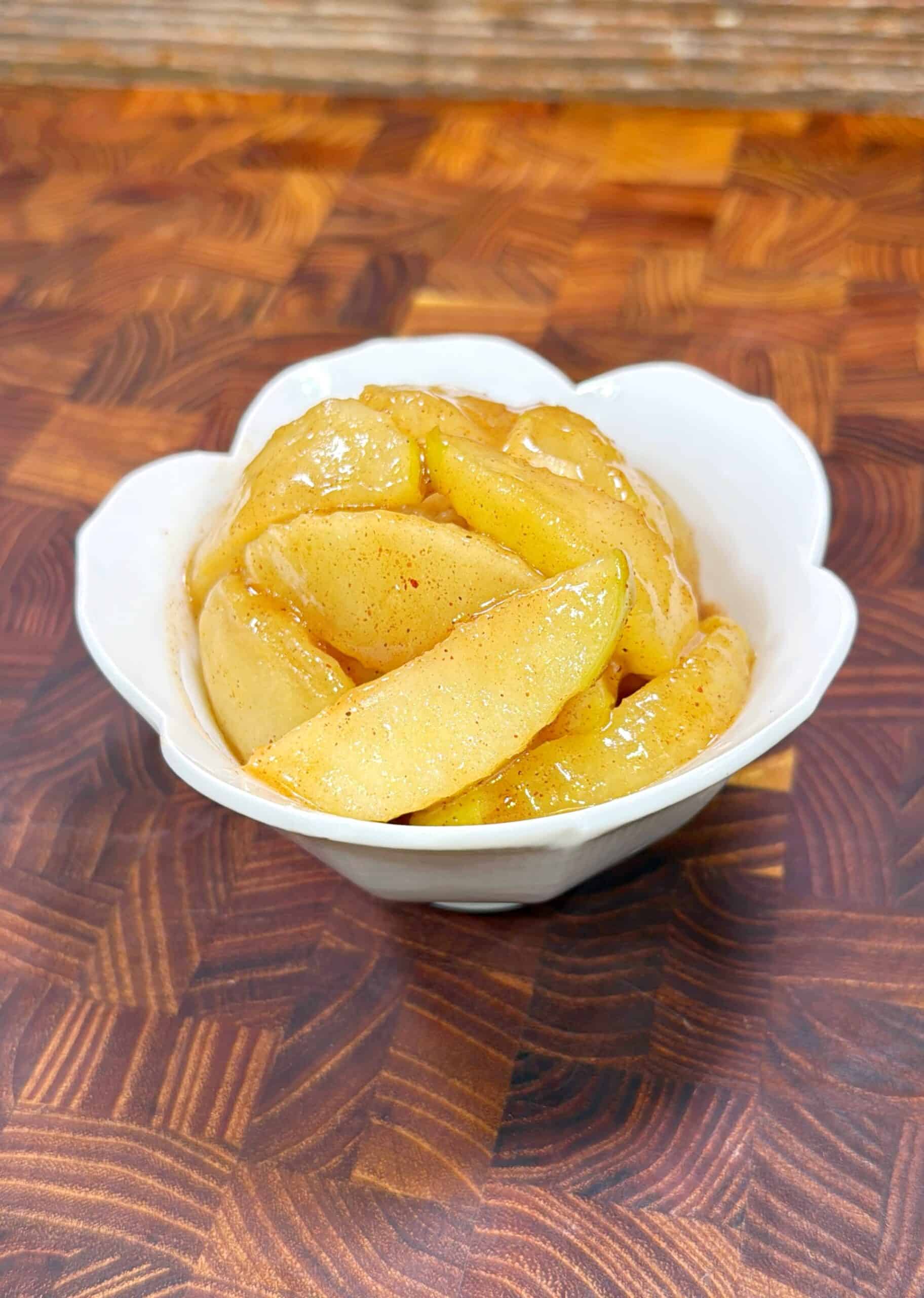 A white bowl filled with glazed, sliced cooked apples sits on a wooden surface with a patterned grain.