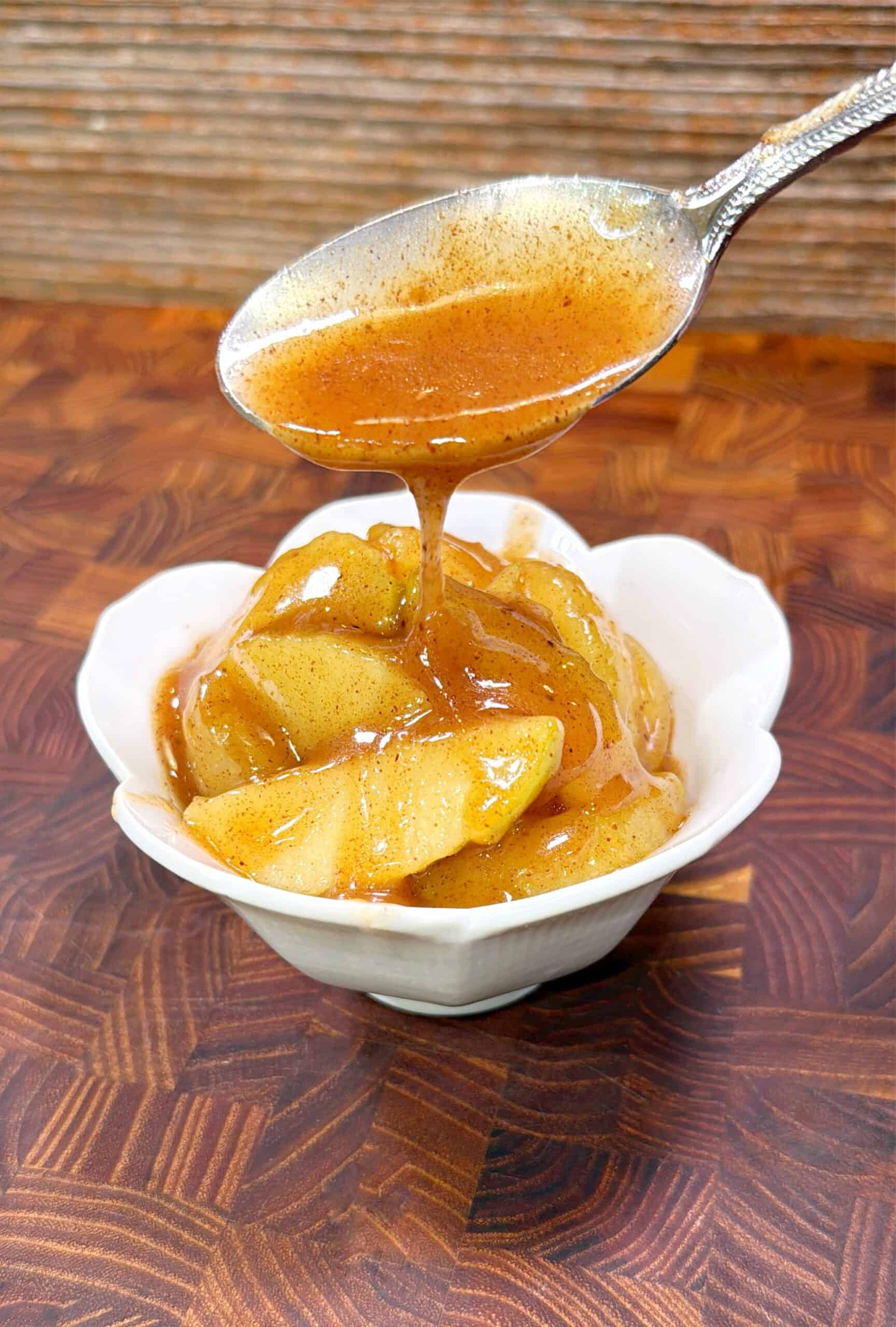 A spoon drizzles cinnamon-spiced sauce over sliced cooked apples in a small white bowl, set on a wooden surface.