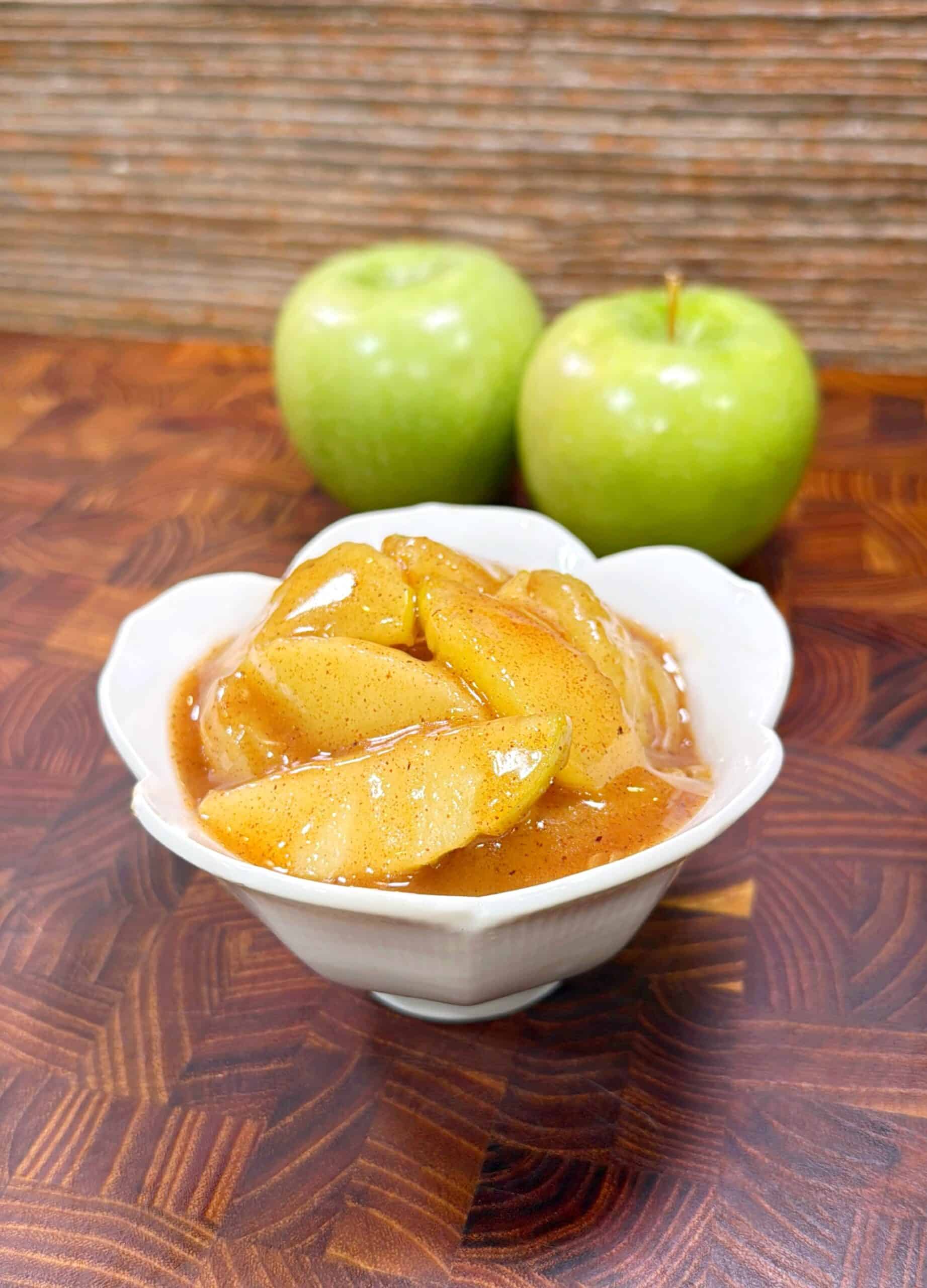 A white bowl filled with cracker barrel apples sits on a wooden surface, with two whole green apples in the background.