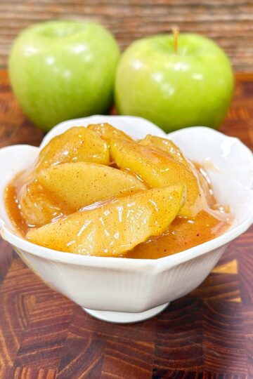 A white bowl filled with glazed, cooked apple slices sits on a wooden surface, with two green apples in the background.