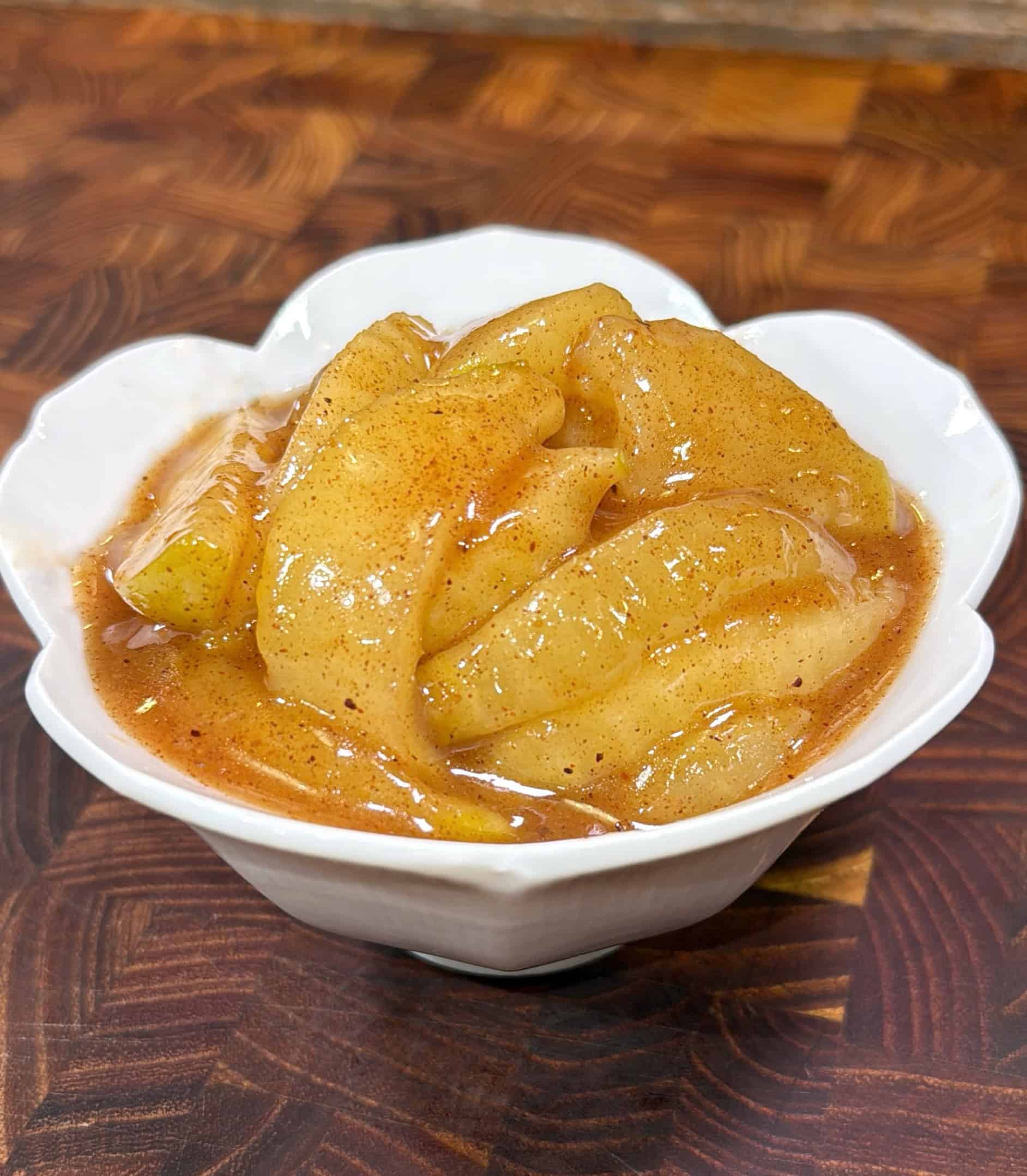 A white, heart-shaped bowl filled with cooked apple slices coated in a glossy cinnamon sauce, set on a patterned wooden surface.
