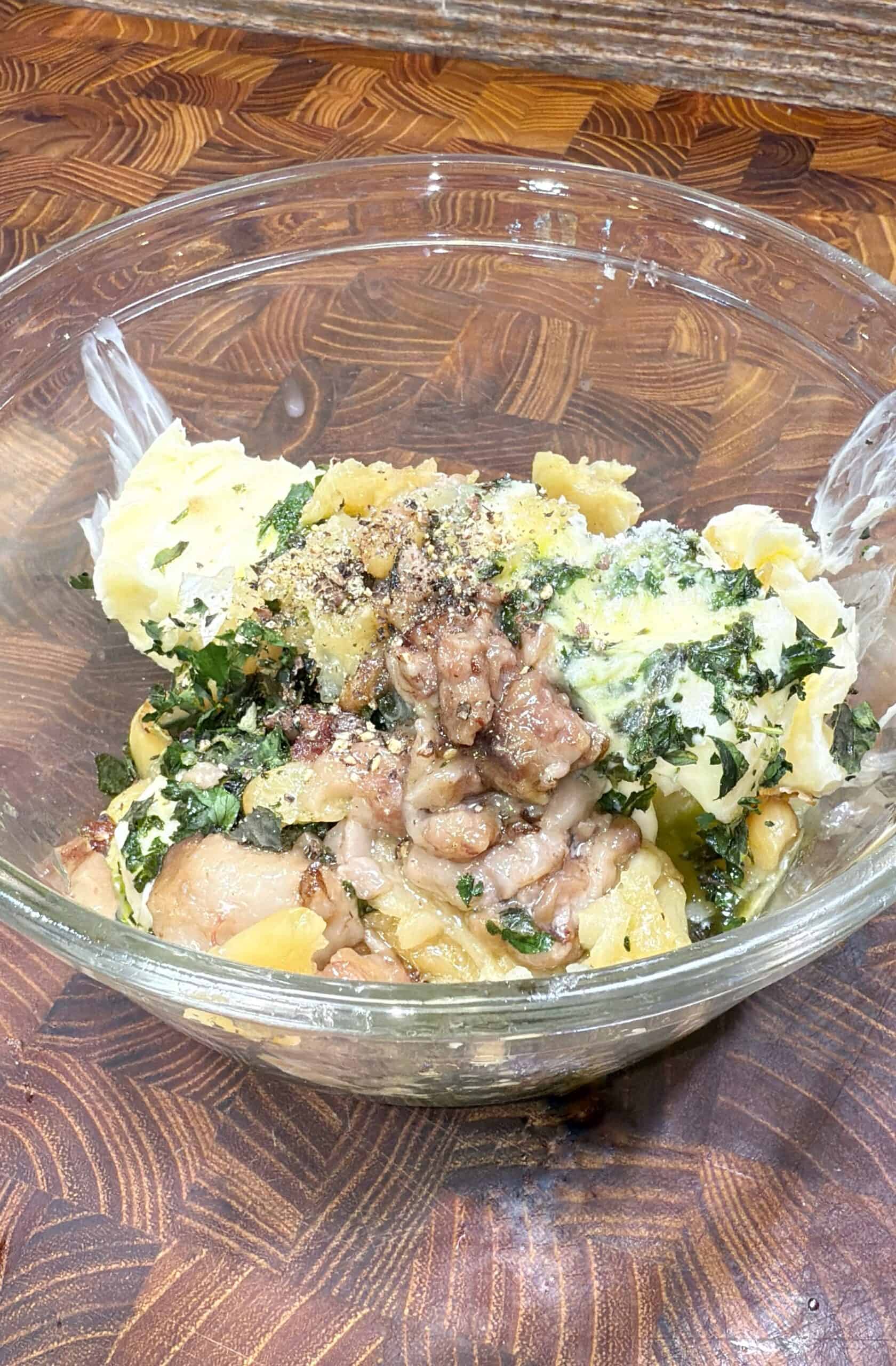A clear glass bowl containing a mixture of mashed potatoes, herbs, ground black pepper, chunks of meat, and rich bone marrow sits on a patterned wooden surface.