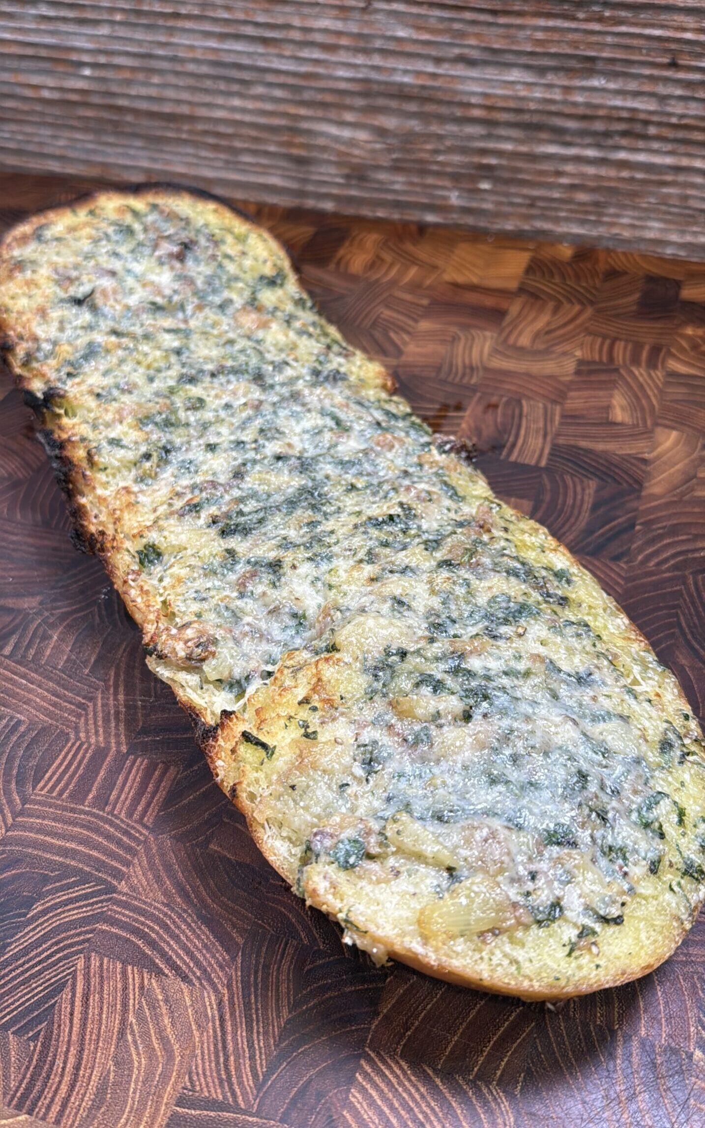 A piece of toasted garlic bread covered with melted herbs and cheese sits on a wooden cutting board.