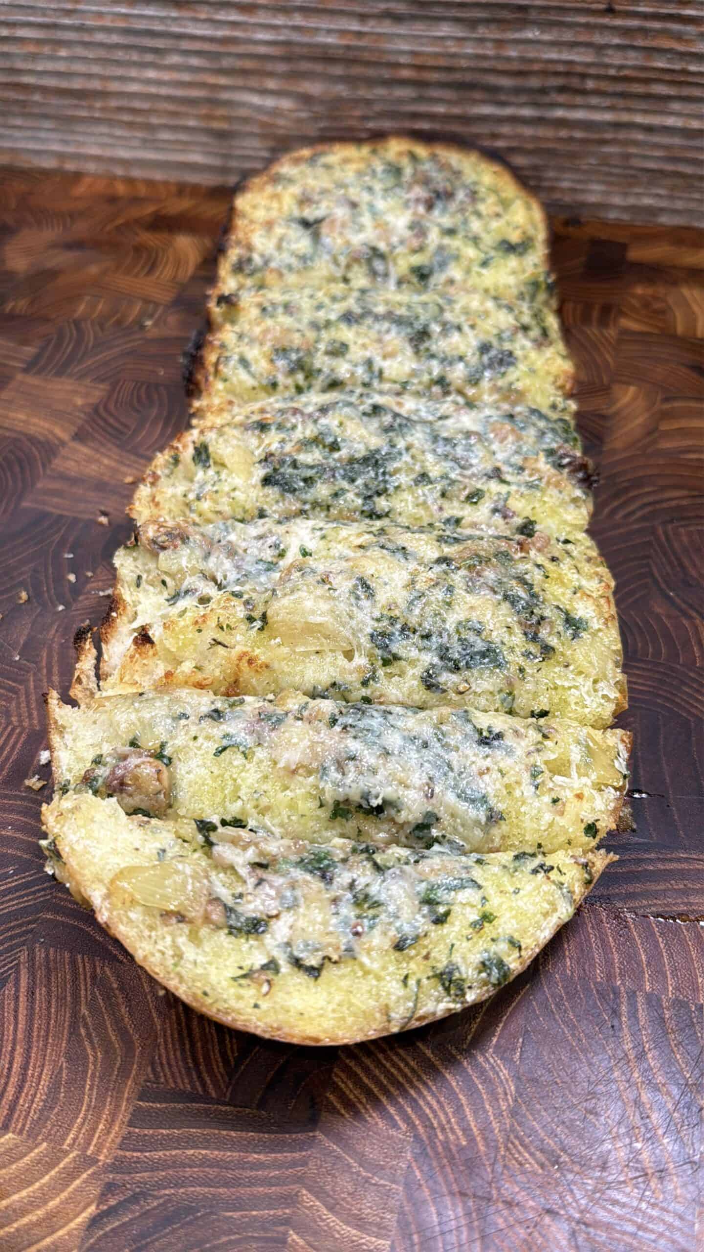 A sliced loaf of garlic bread topped with herbs and melted cheese is displayed on a wooden cutting board with a patterned surface.