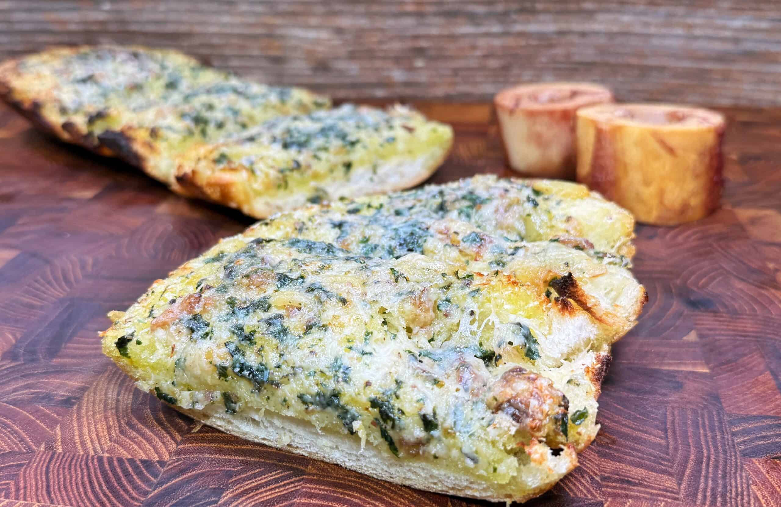 A close-up of rectangular slices of cheesy garlic bread topped with herbs, set on a wooden surface. Two small, round pieces of bread are blurred in the background.