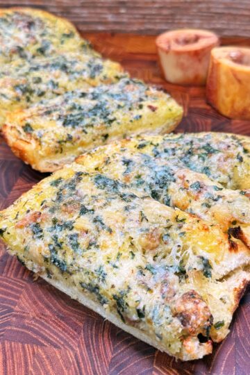 Sliced pieces of toasted bread topped with a green herb and cheese mixture are arranged on a wooden board, with two roasted bone marrow pieces in the background.