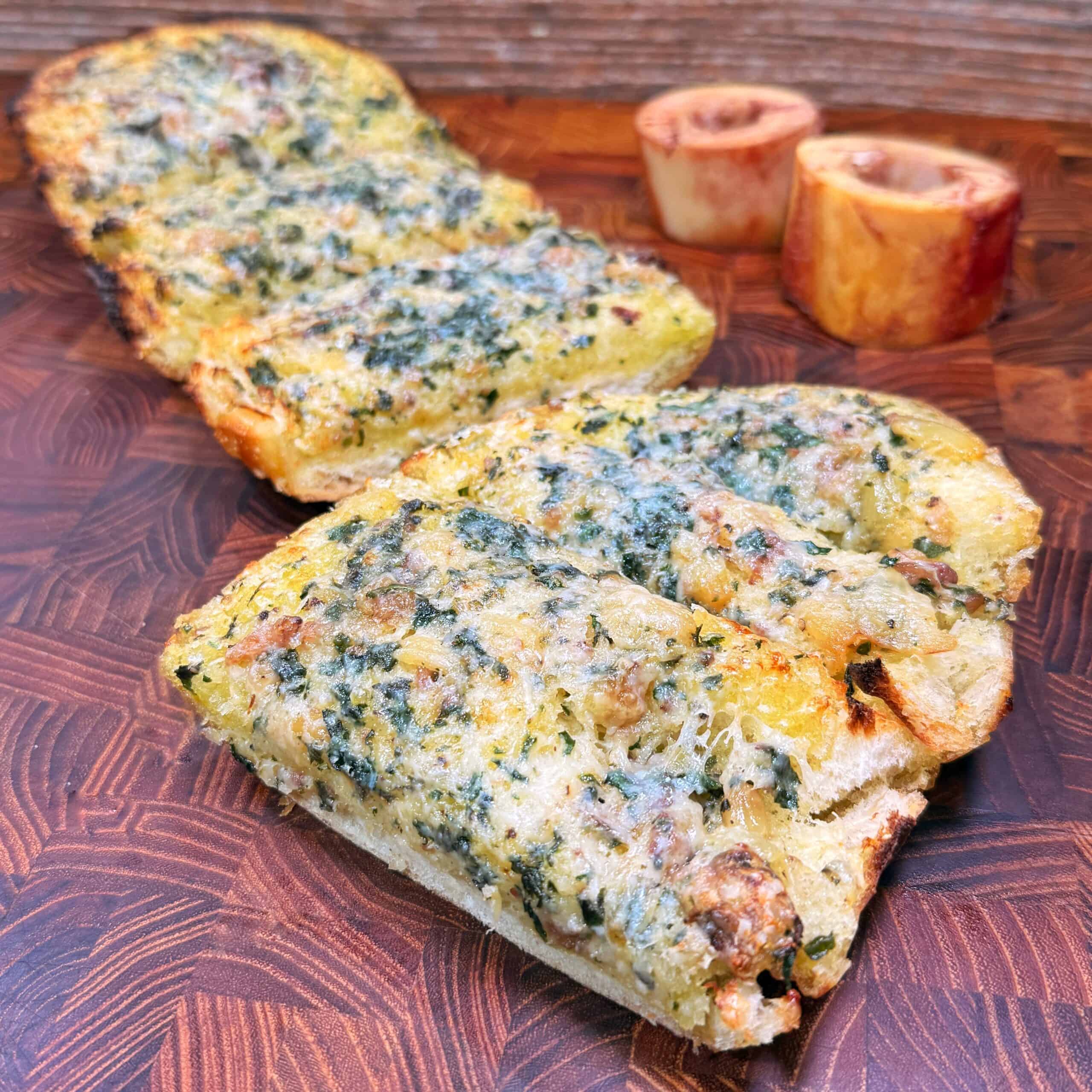 Sliced pieces of bone marrow garlic  bread topped with a green herb and cheese mixture are arranged on a wooden board, with two roasted bone marrow pieces in the background.
