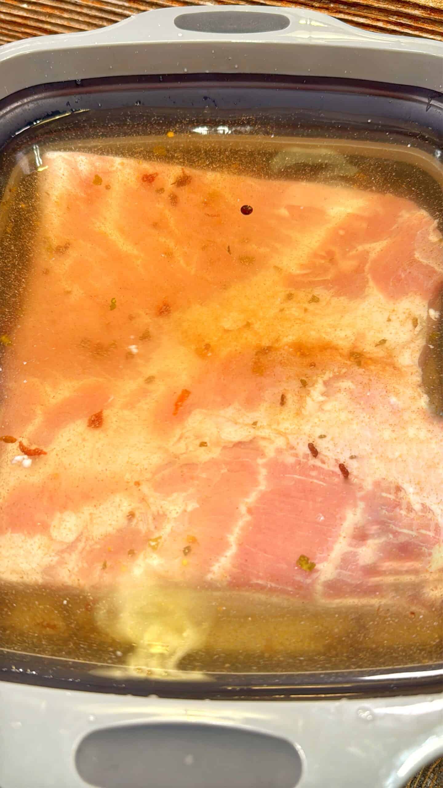 Raw bacon slices submerged in a clear brine with visible spices and seasonings floating in the liquid, inside a rectangular container.