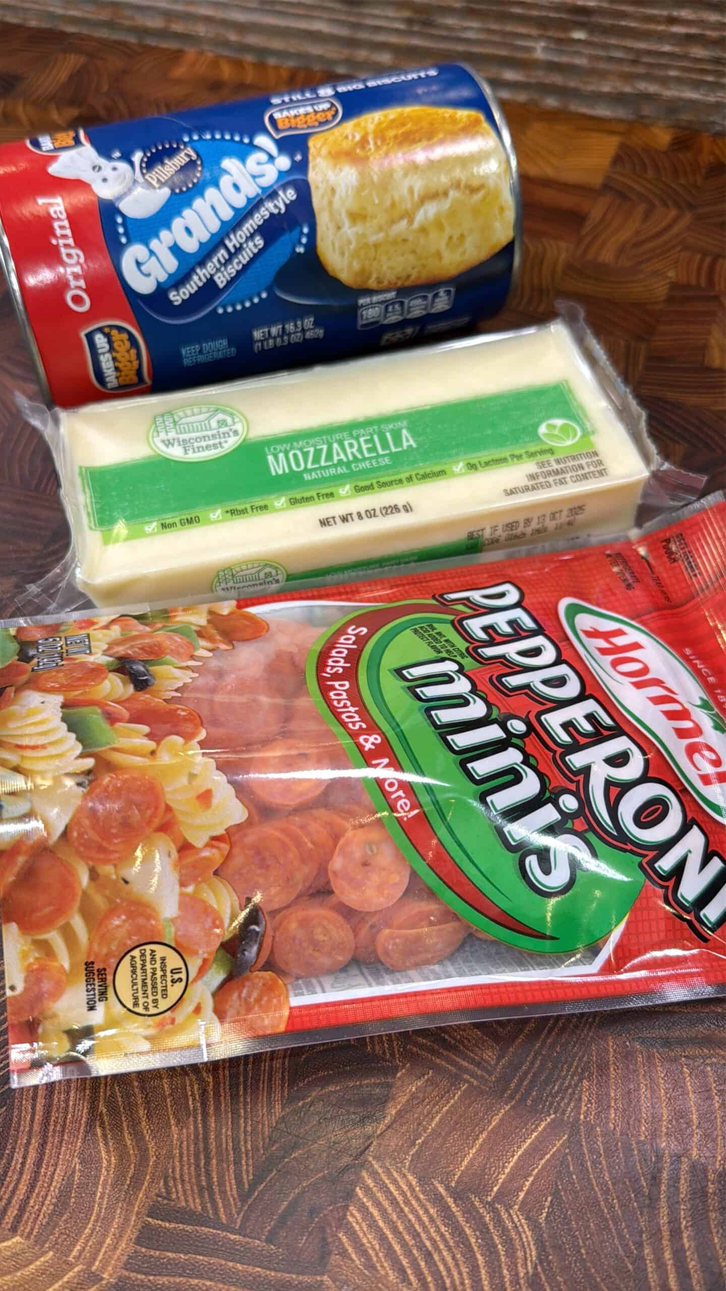 A package of Grands! Southern Homestyle biscuits, a block of mozzarella cheese, and a bag of Hormel pepperoni minis on a wooden surface.