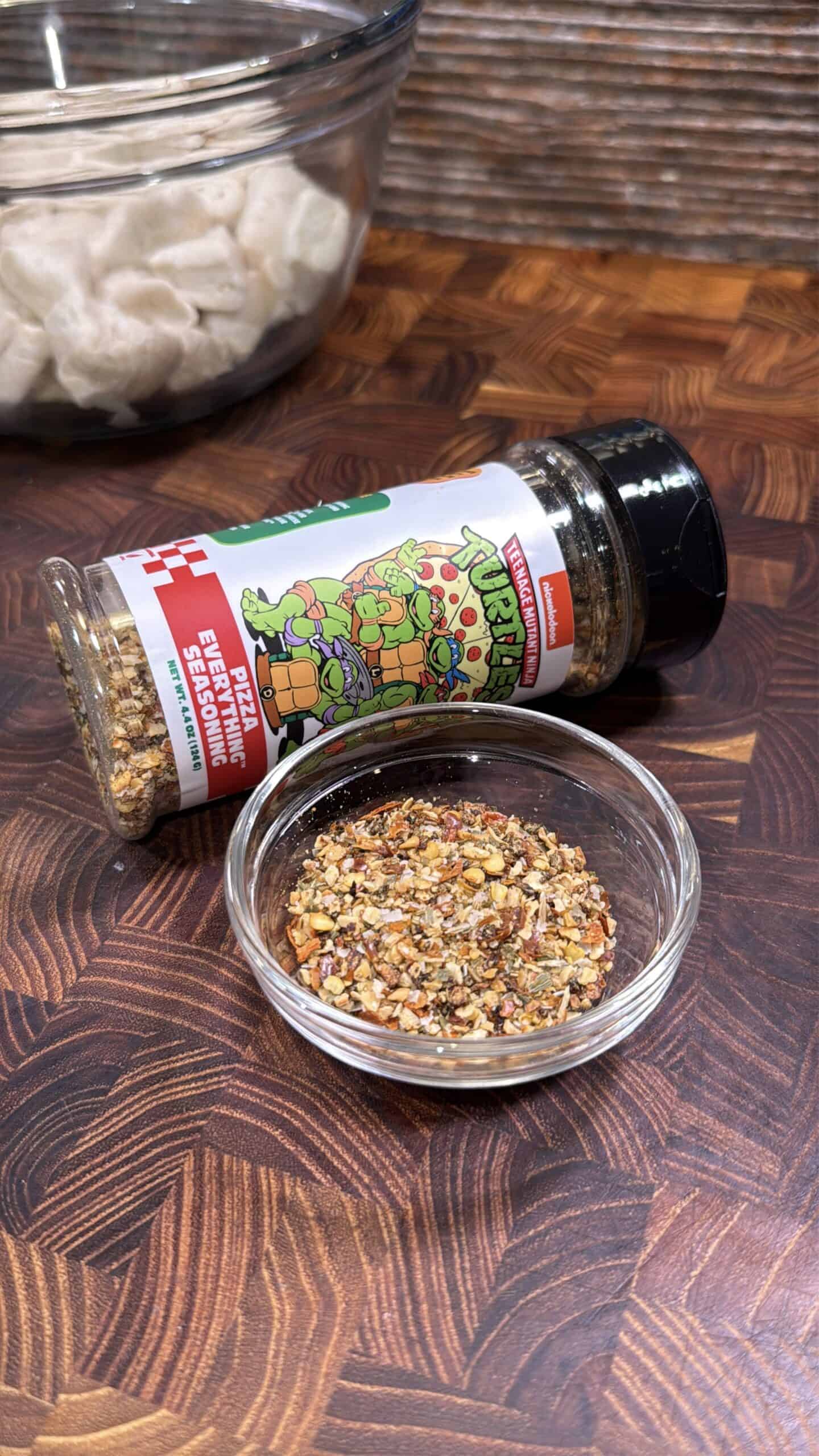 A glass bowl and a bottle of Teenage Mutant Ninja Turtles extra pizza seasoning are on a wooden surface. The bowl contains some of the seasoning, and a bowl of uncooked dumplings is in the background.