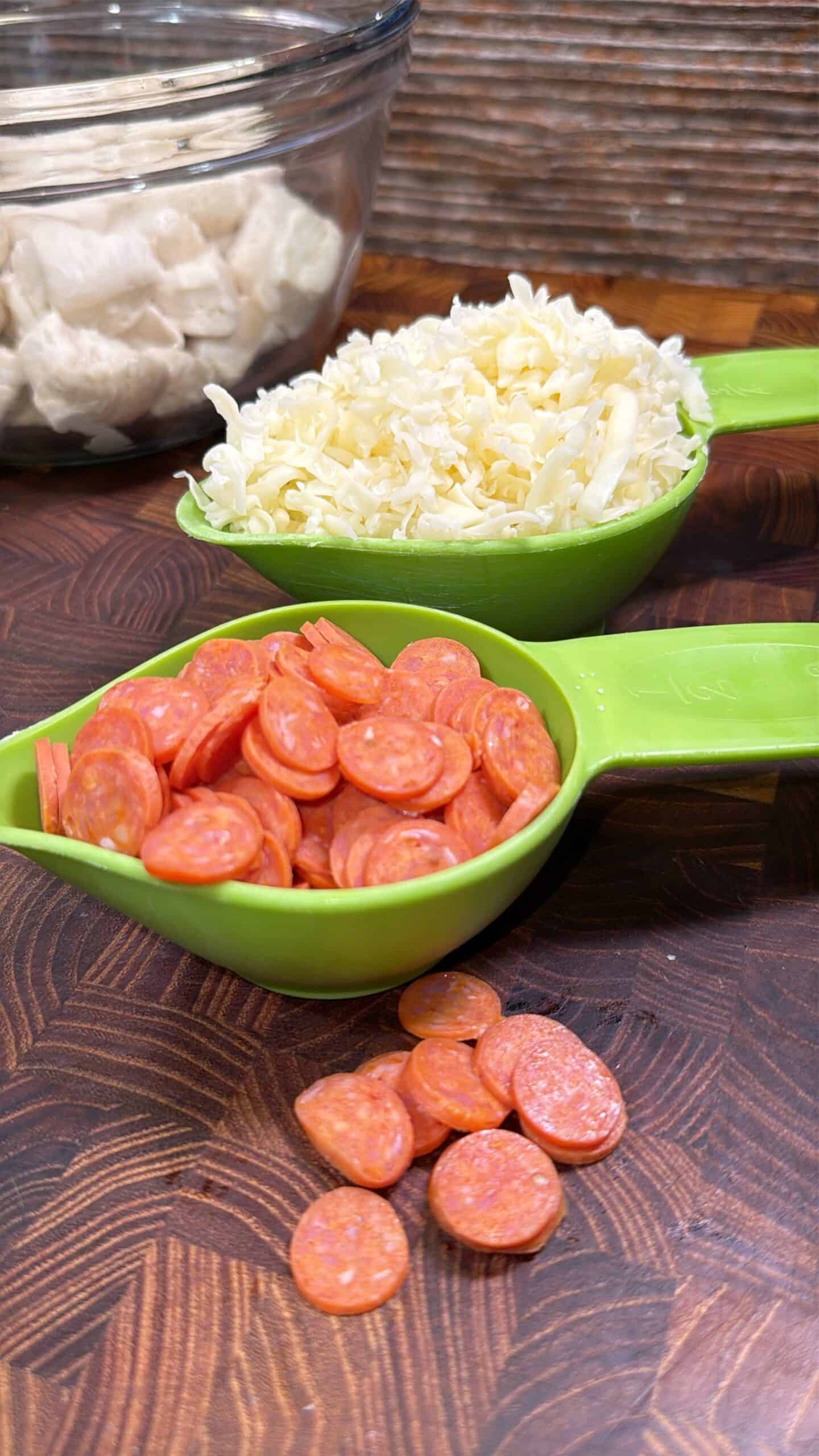 Two green measuring cups hold shredded mozzarella cheese and mini pepperoni slices. Some pepperoni slices are scattered on a wooden surface. In the background, a glass bowl contains dough pieces.