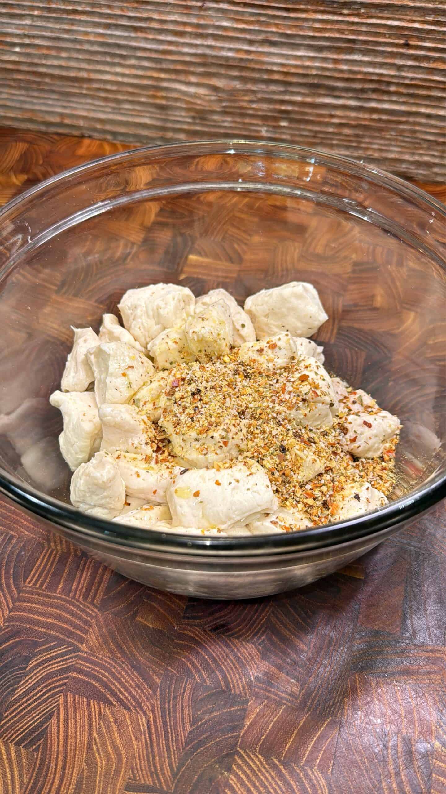 Glass bowl filled with raw, cubed chicken pieces topped with a mix of colorful spices, sitting on a patterned wooden surface.