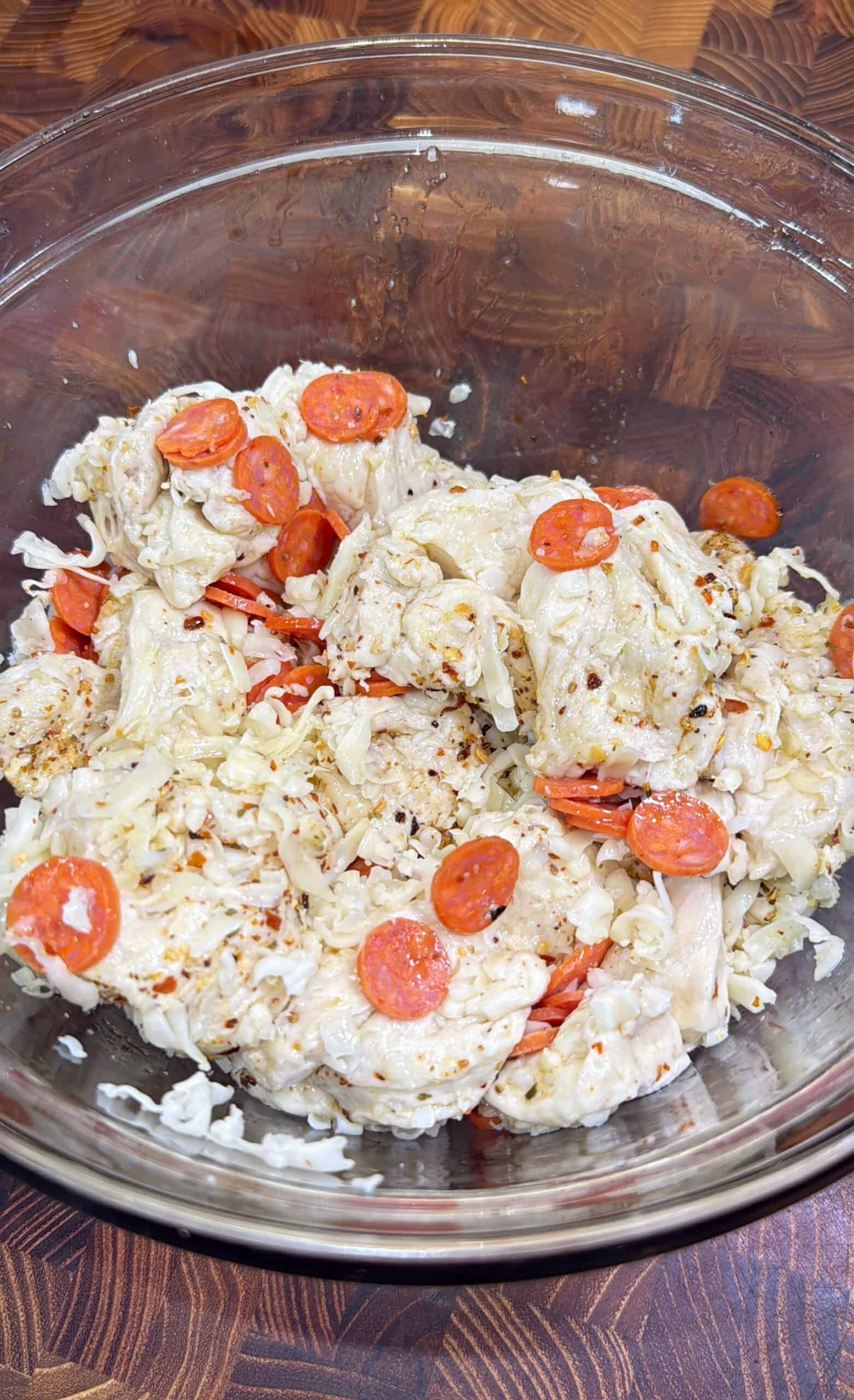 A glass bowl filled with a mixture of shredded chicken, mini pepperoni slices, mozzarella cheese, and Italian seasoning, sitting on a wooden surface.