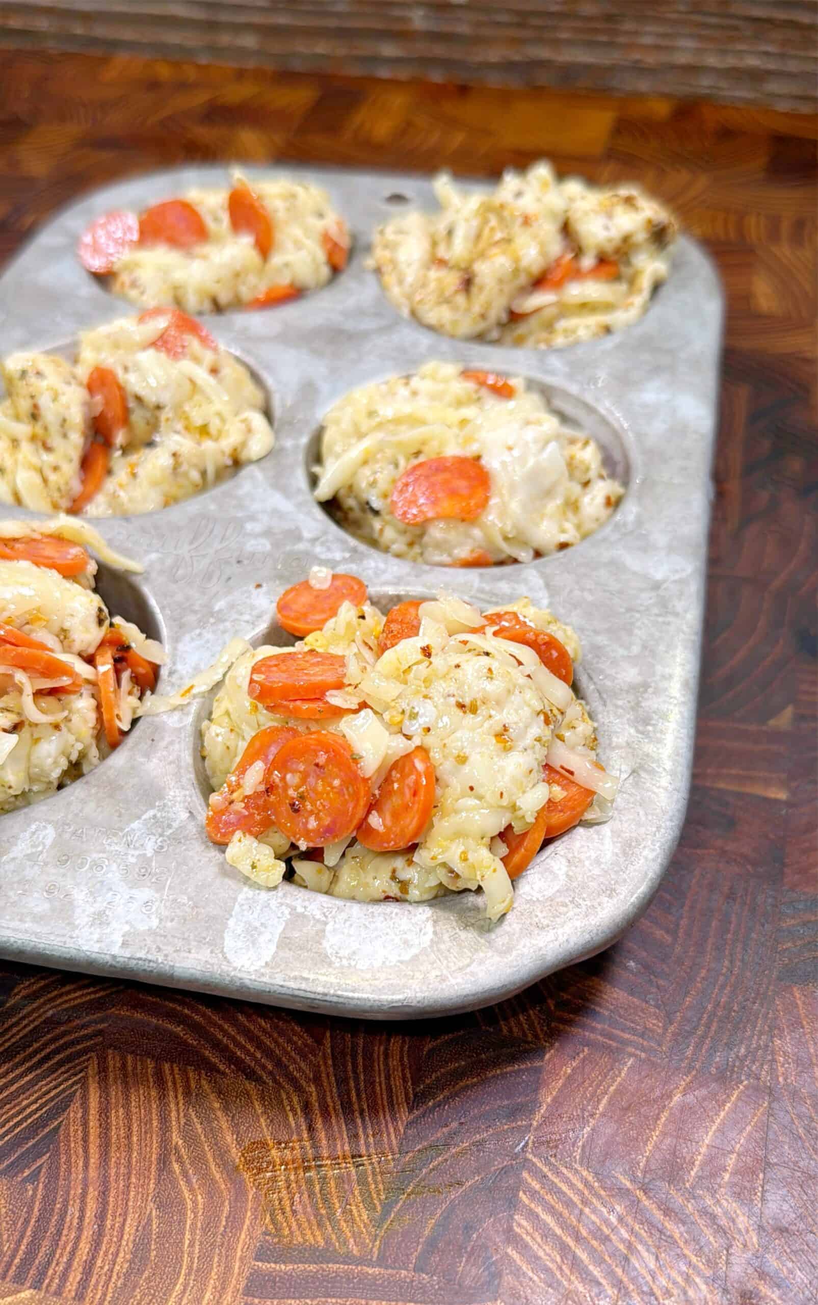 A metal muffin tin holding several baked muffin-shaped pizza bites with visible pepperoni slices, melted cheese, and herbs, resting on a wooden surface.