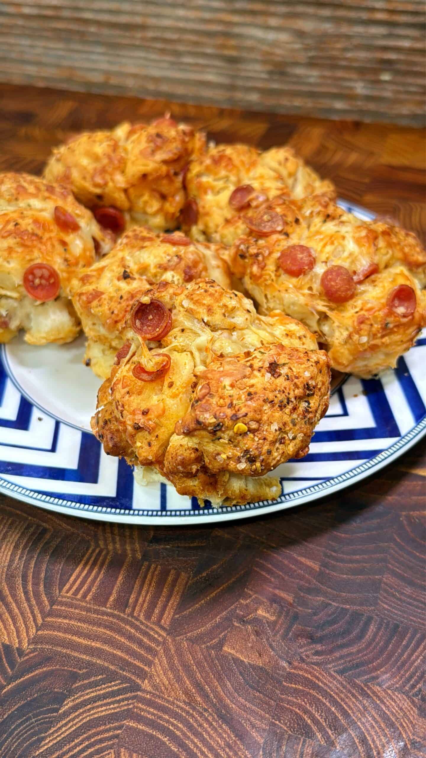 A plate filled with golden-brown, cheesy bread rolls topped with small pepperoni slices, arranged on a blue and white patterned plate, sitting on a wooden surface.