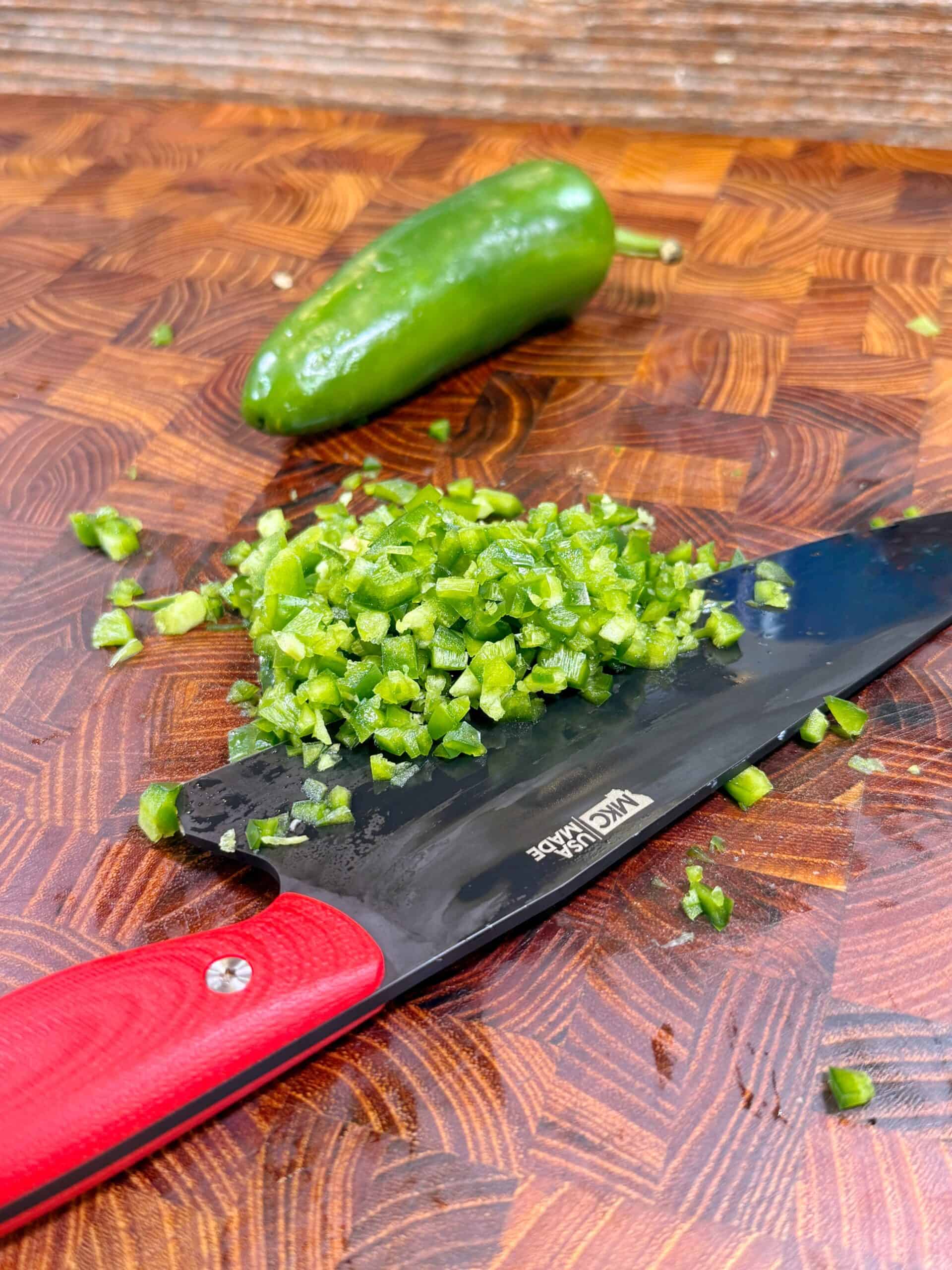 A knife with a red handle holds chopped green jalapeño peppers on a wooden cutting board, with a whole jalapeño pepper in the background.
