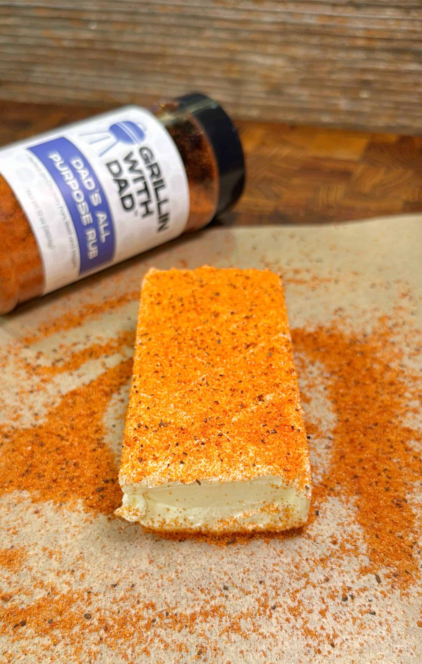 A rectangular block of cream cheese coated with orange seasoning sits on parchment paper, with a bottle of Dads All Purpose Rub grill seasoning lying behind it. Seasoning is sprinkled around the cheese.