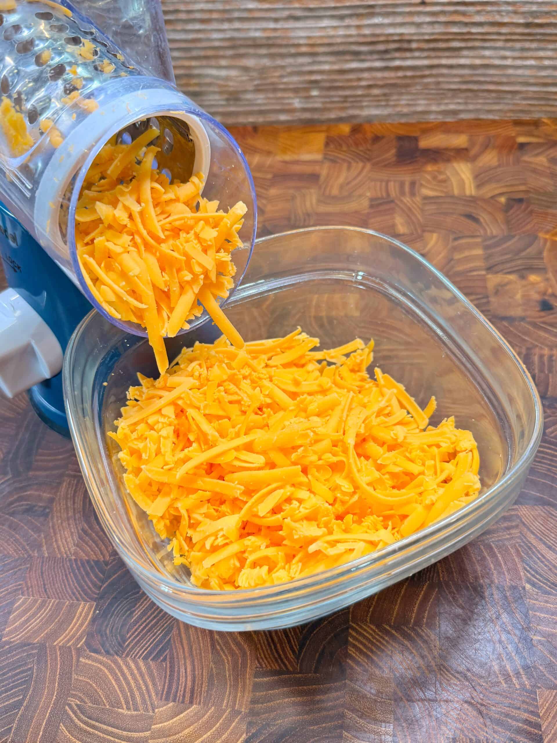 Shredded orange cheese falling from a rotary grater into a clear glass bowl, placed on a wooden countertop with a textured background.