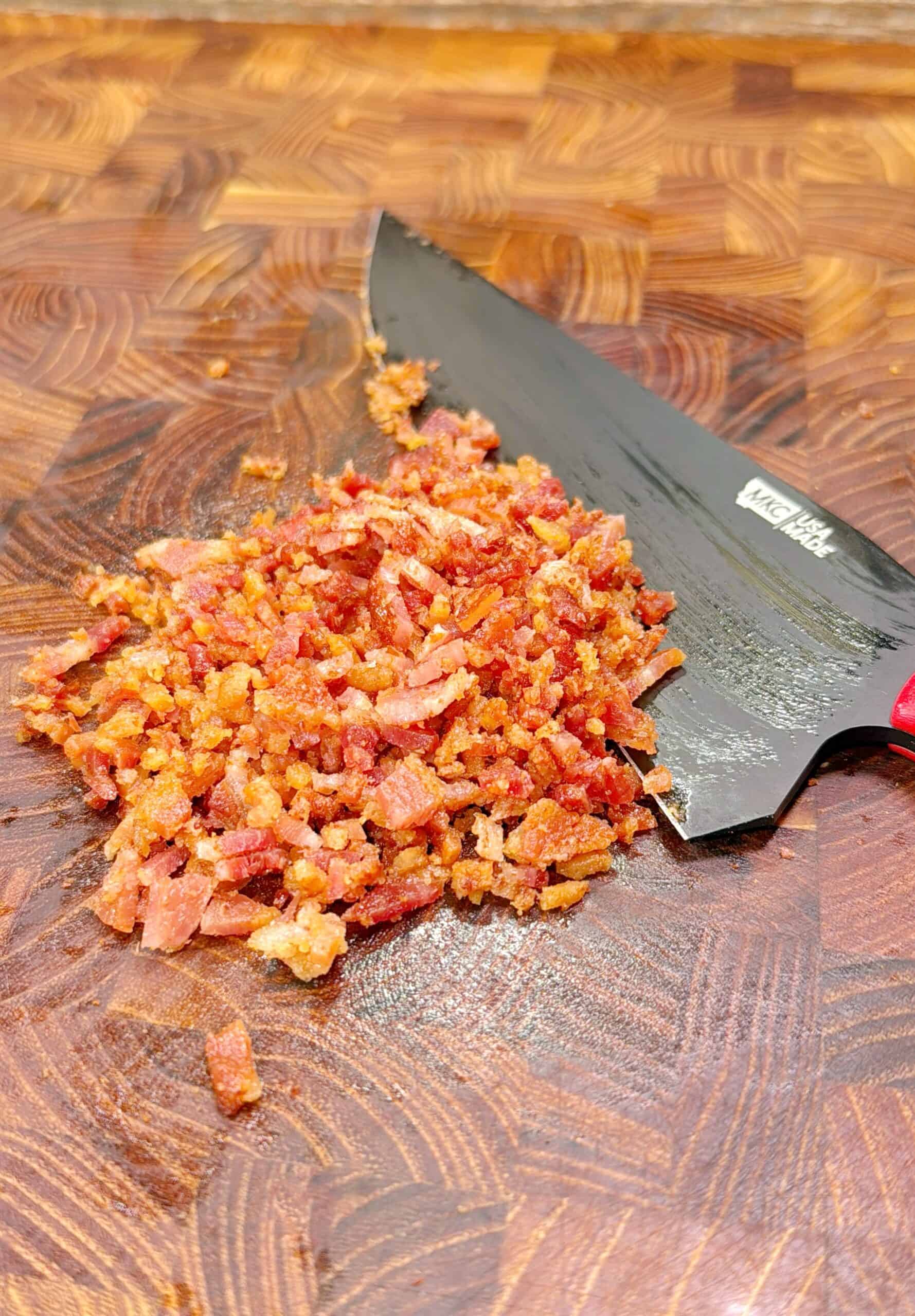 Chopped crispy bacon pieces on a wooden cutting board next to a black knife with a red handle.