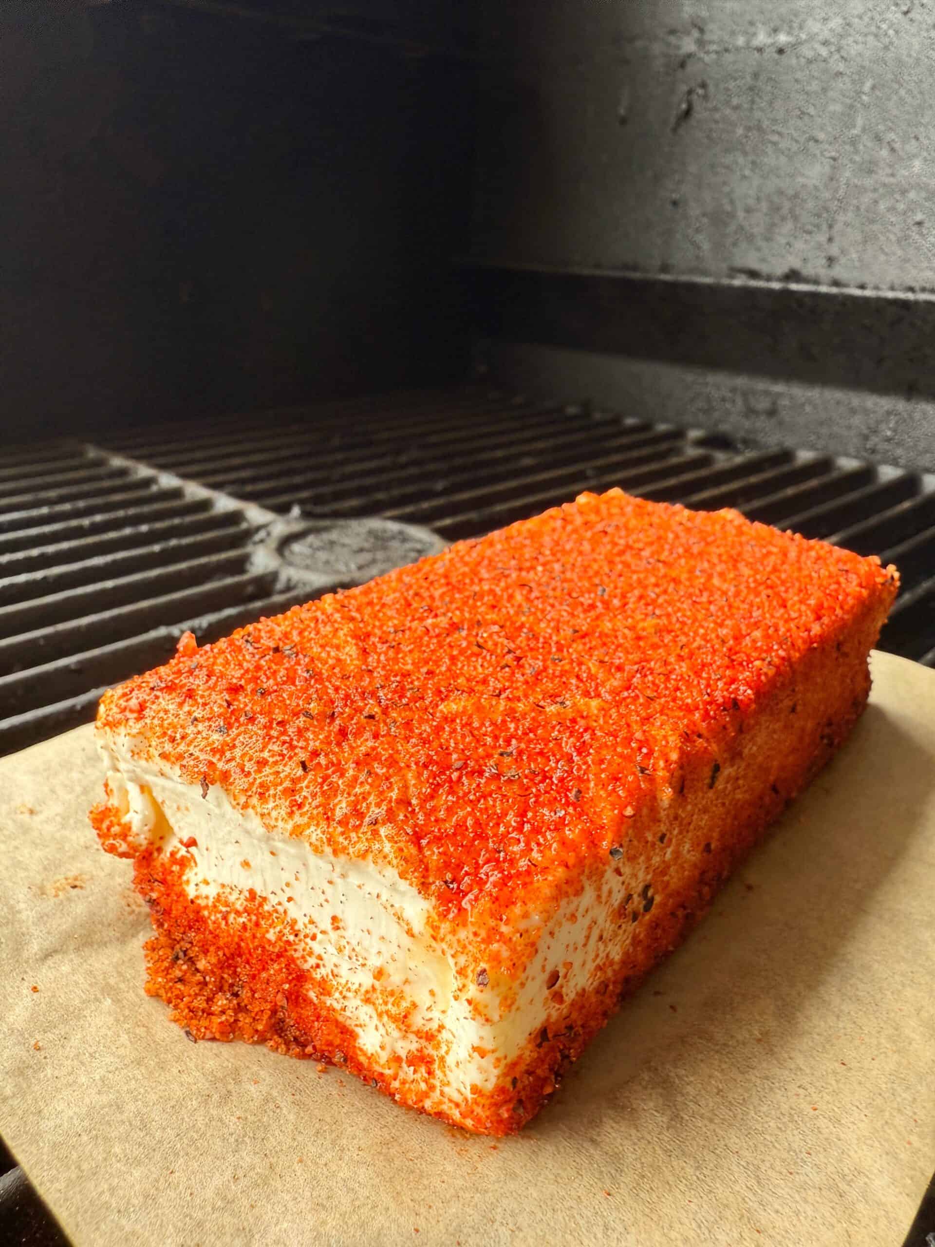 A rectangular block of white cheese, coated in red seasoning, sits on parchment paper on a grill with dark metal grates.