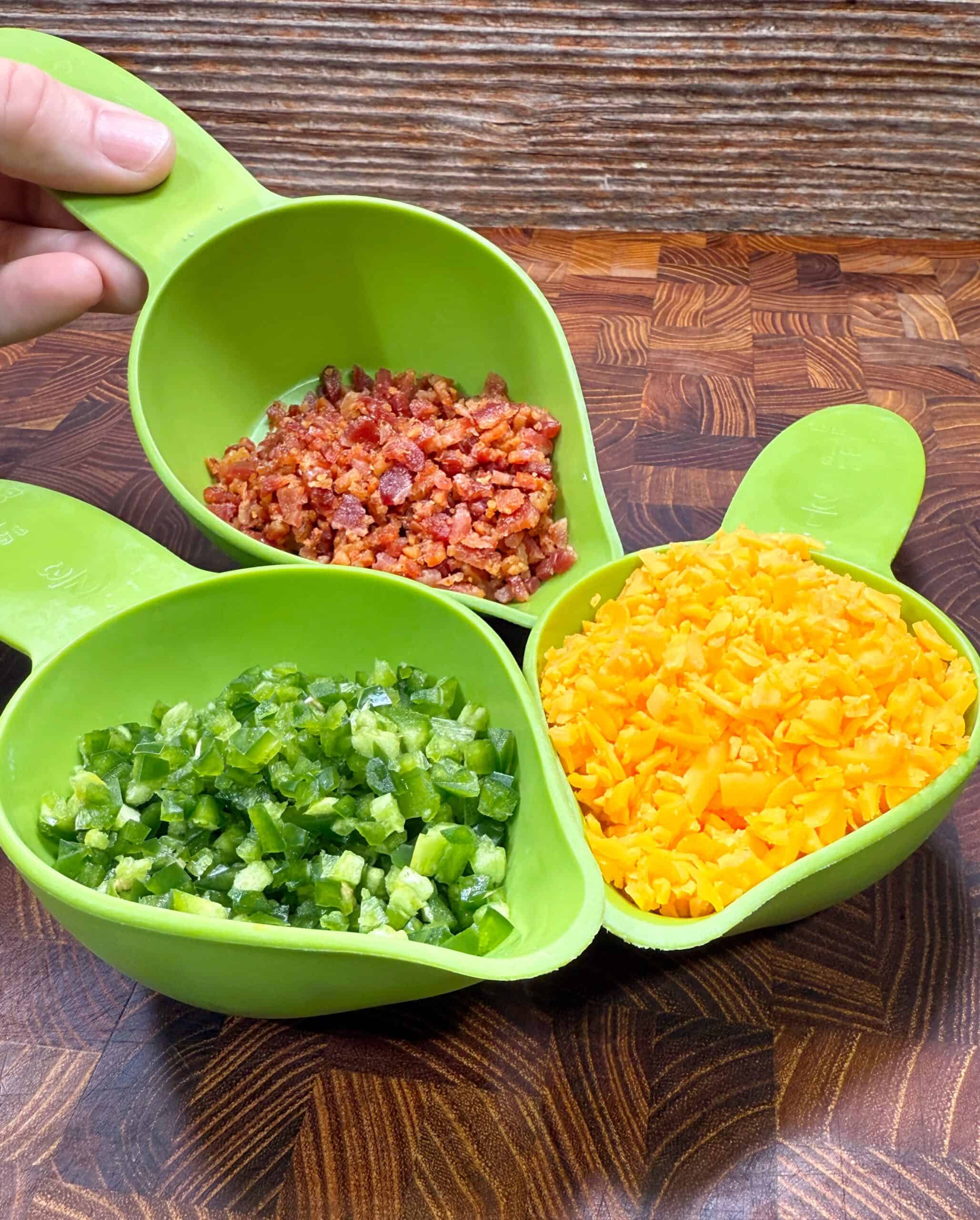 Three green measuring cups filled with diced cooked bacon, chopped green jalapeños, and shredded yellow cheddar cheese are arranged on a wood countertop. A hand holds the cup with bacon.