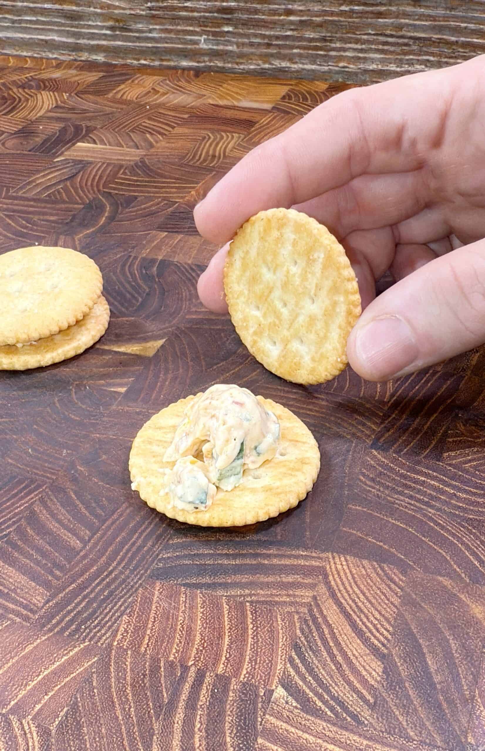 A hand holds a round cracker above another cracker topped with a creamy spread on a wooden surface. A small stack of plain crackers sits in the background.