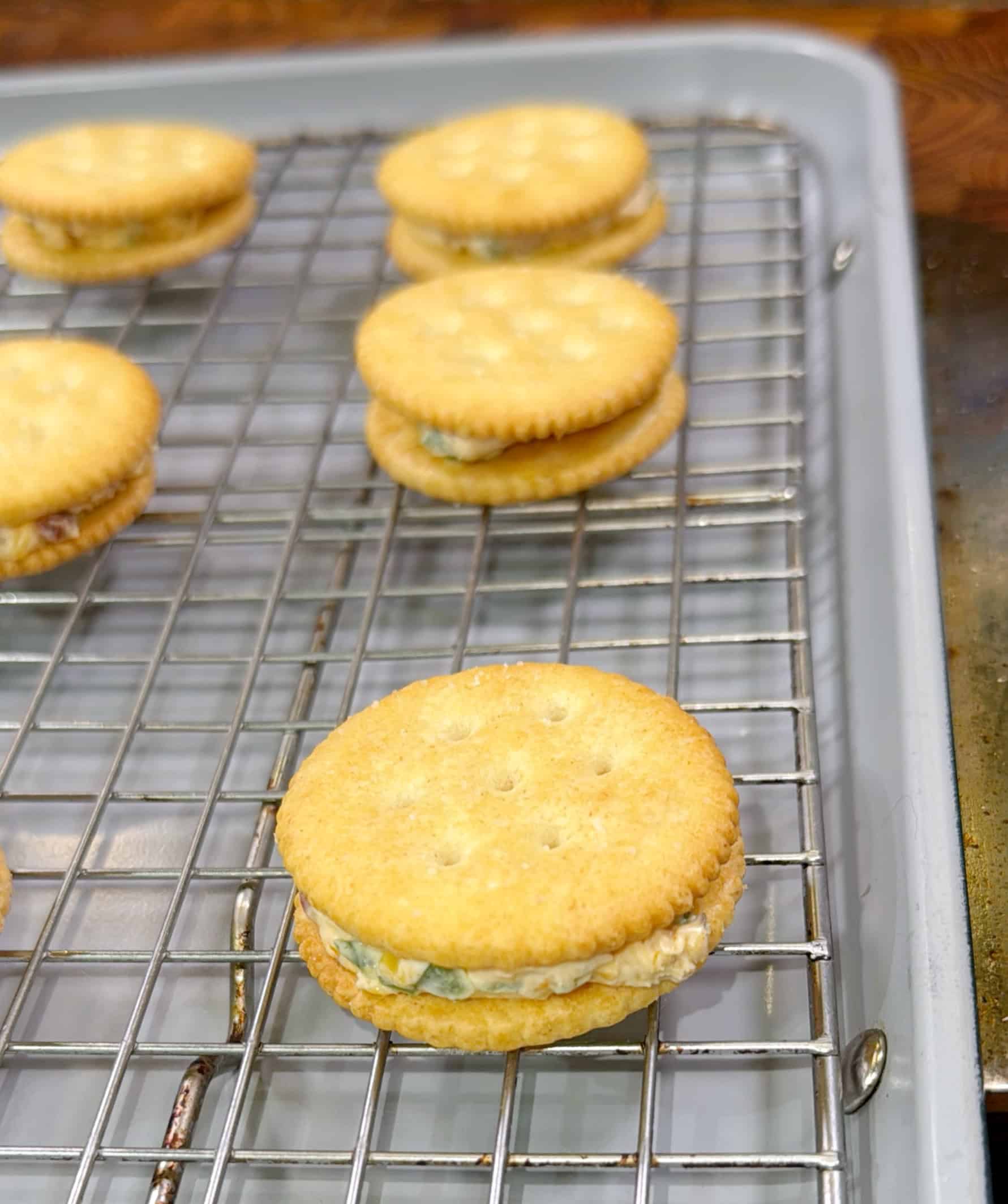 Several sandwich crackers with a creamy filling are placed on a wire cooling rack over a baking sheet. The crackers are round and golden brown, and the filling appears to have herbs mixed in.