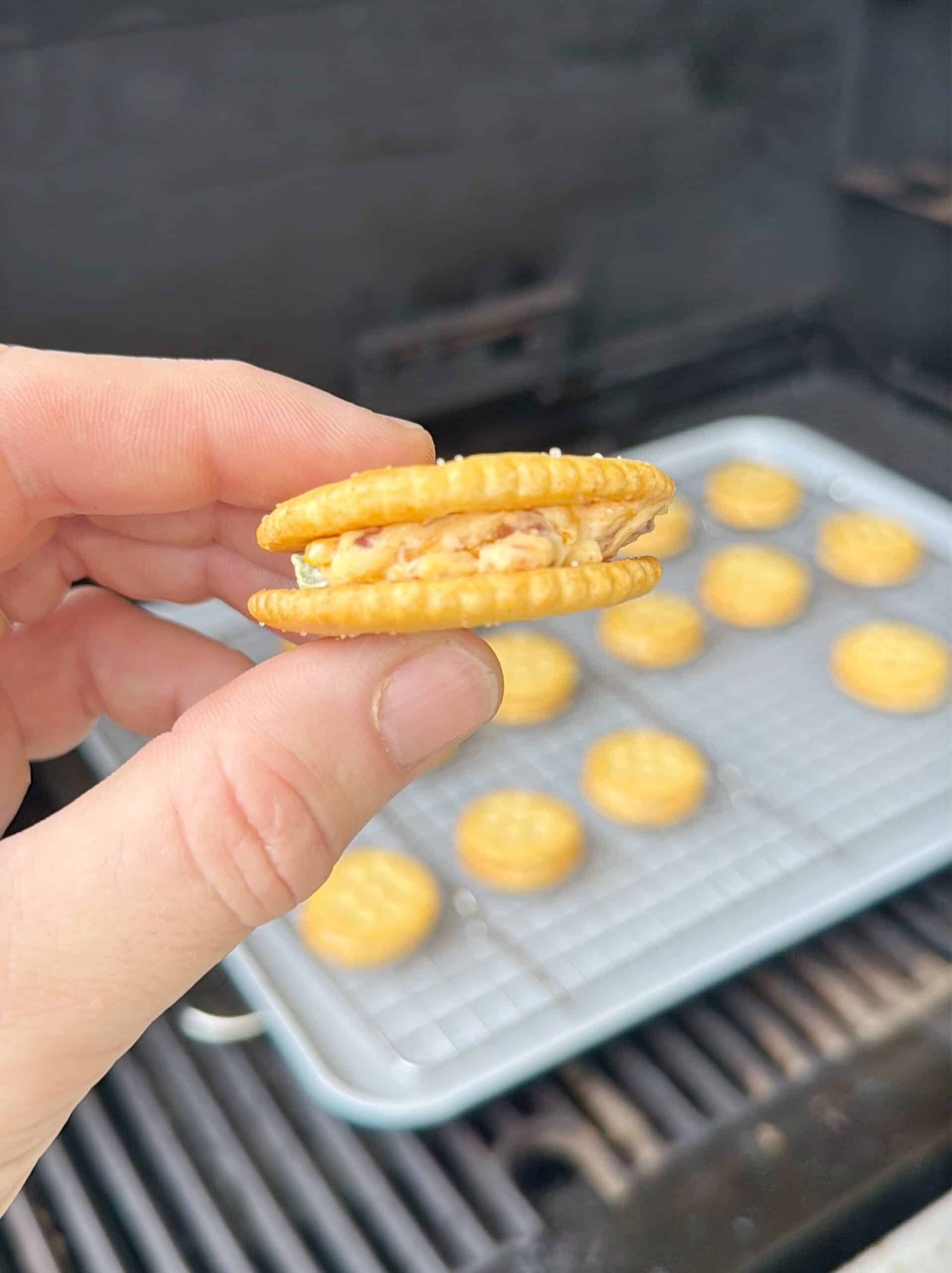 A hand holds a sandwich cracker with a creamy filling. In the background, more sandwich crackers are arranged on a wire rack over a baking sheet.